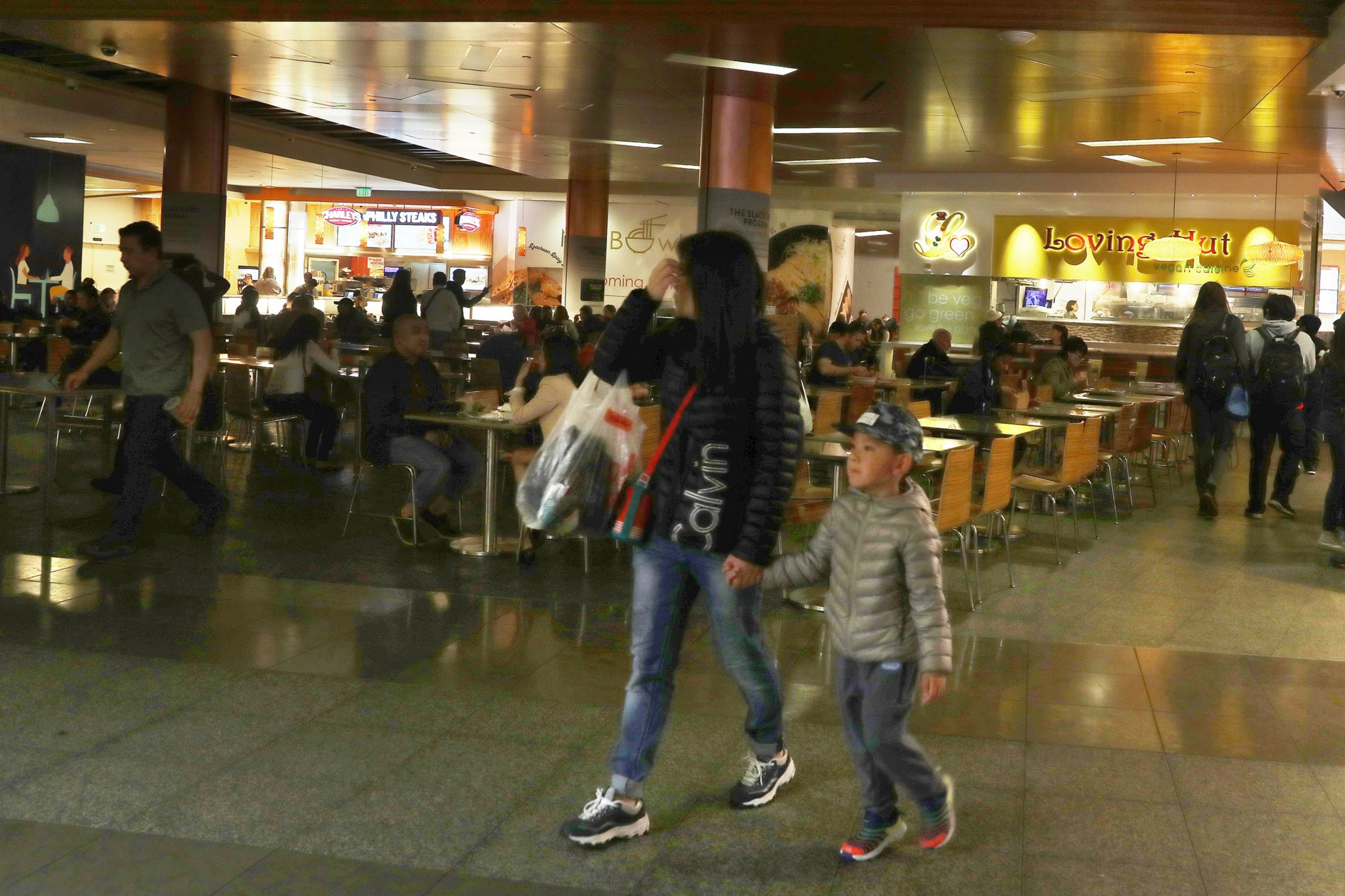 2 people walk through a busy food court with low lighting in background