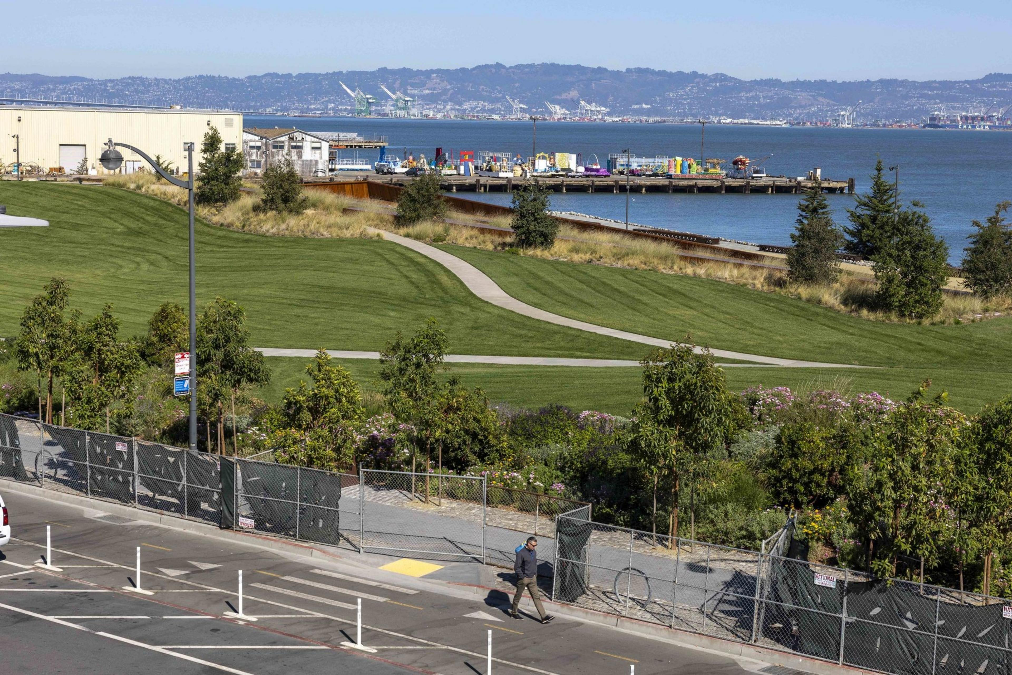 A green park by the waterfront features a winding path. Near the shore are industrial buildings and colorful containers. A person walks on the pavement.
