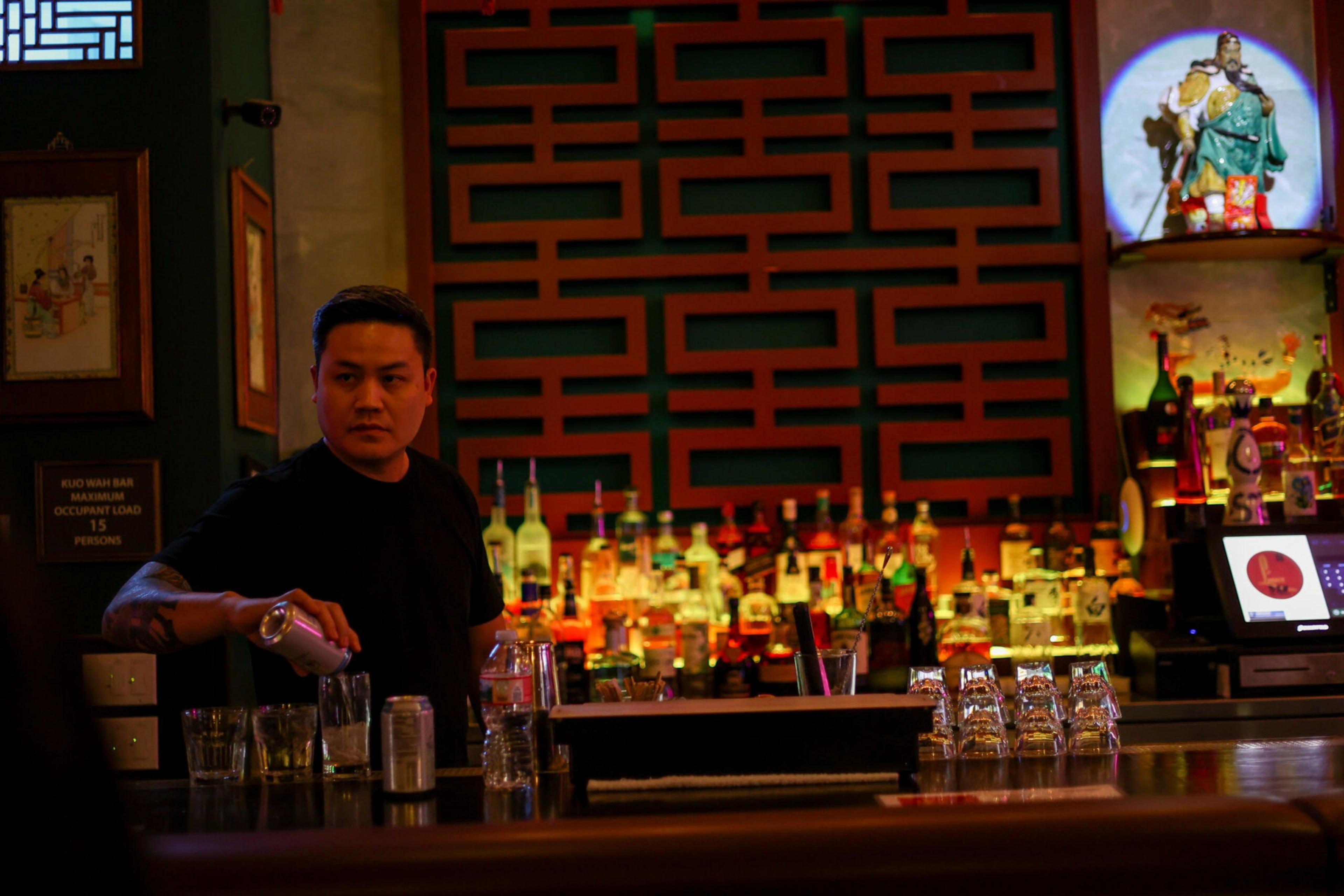 A bartender pours a drink behind a dimly lit bar with many bottles, glasses, and a decorative wall and figurine in the background.