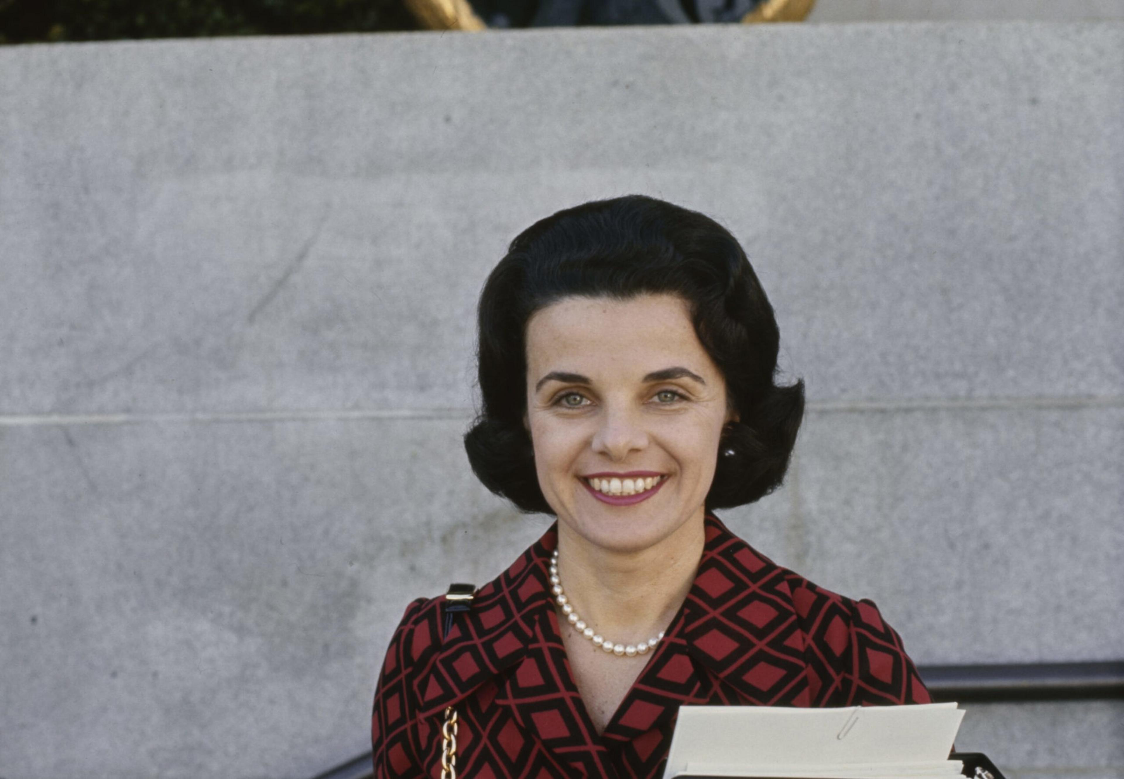 A woman with dark hair styled in waves wears a red and black diamond-patterned coat, pearl necklace, and smiles while holding papers.