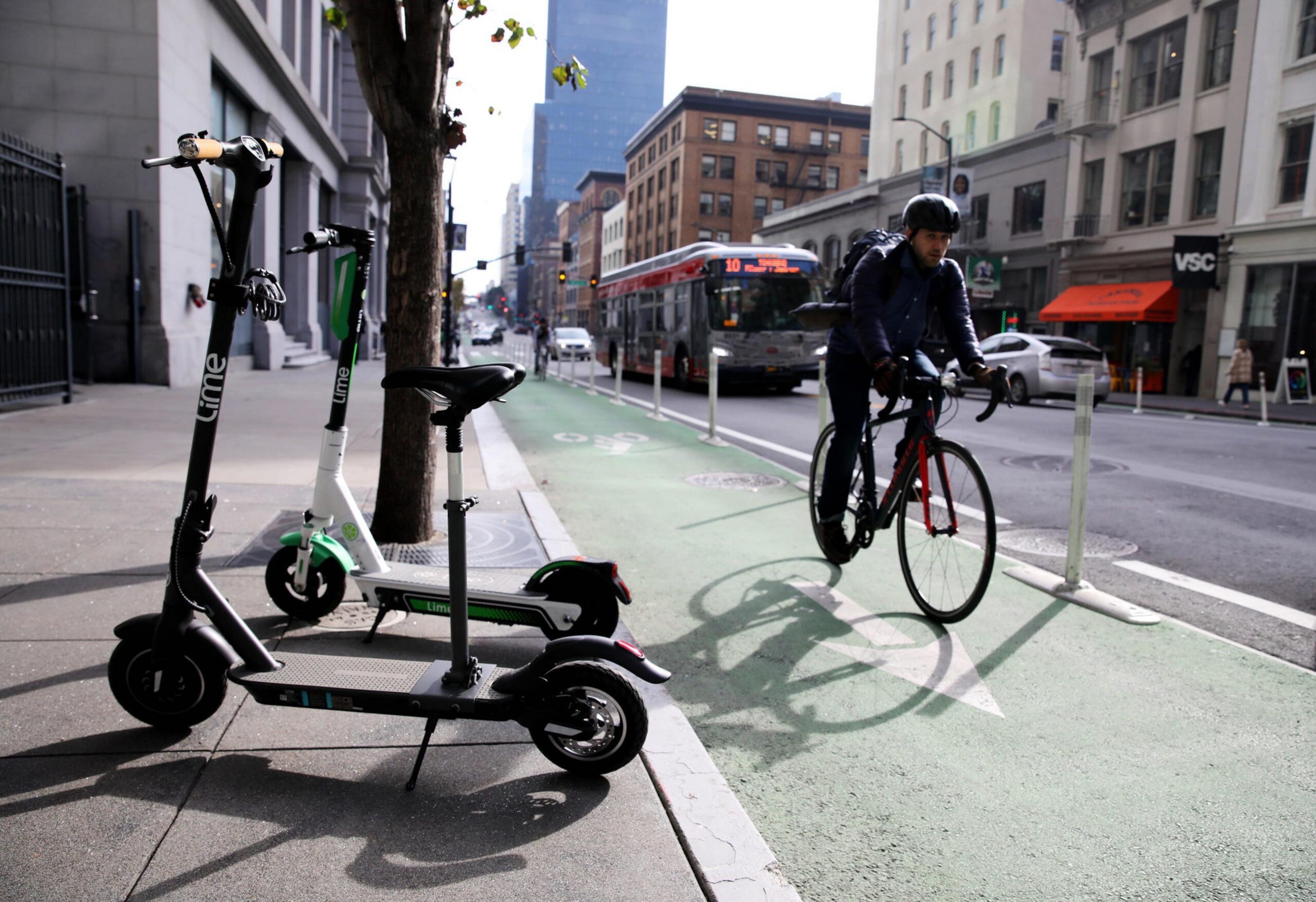 Two electric scooters are parked on the sidewalk next to a green bike lane, where a person wearing a helmet rides a bike past a city bus.