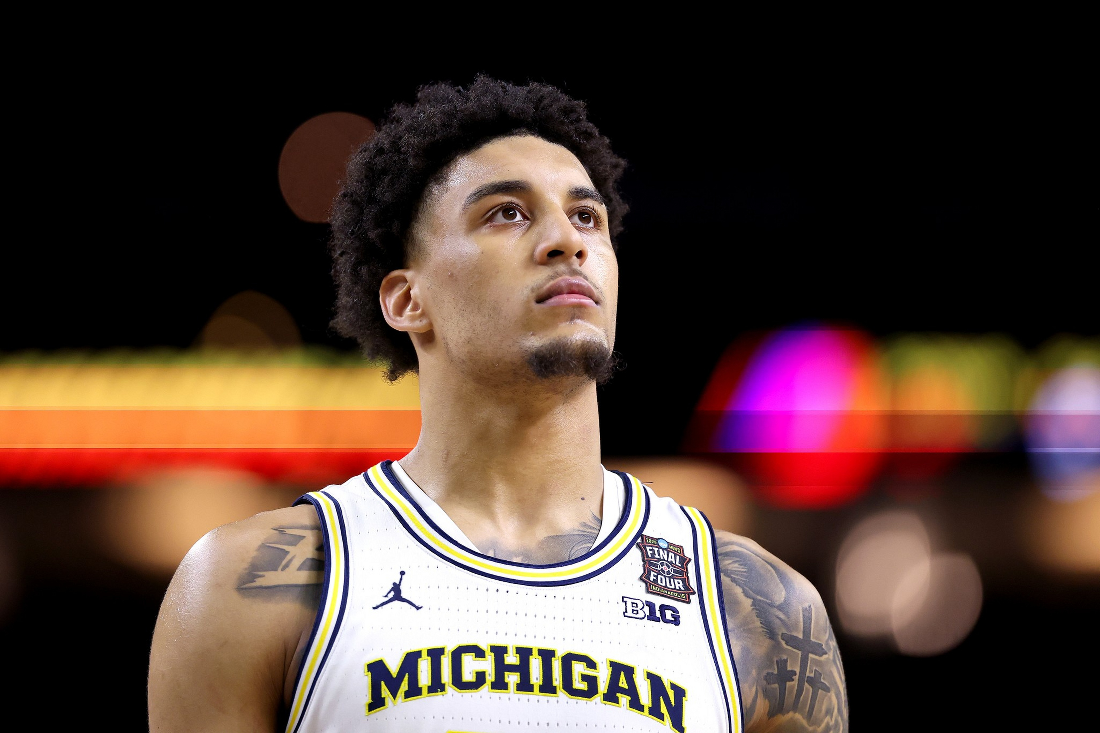 A basketball player in a Michigan jersey looks upward with a focused expression against a blurred, colorful background.