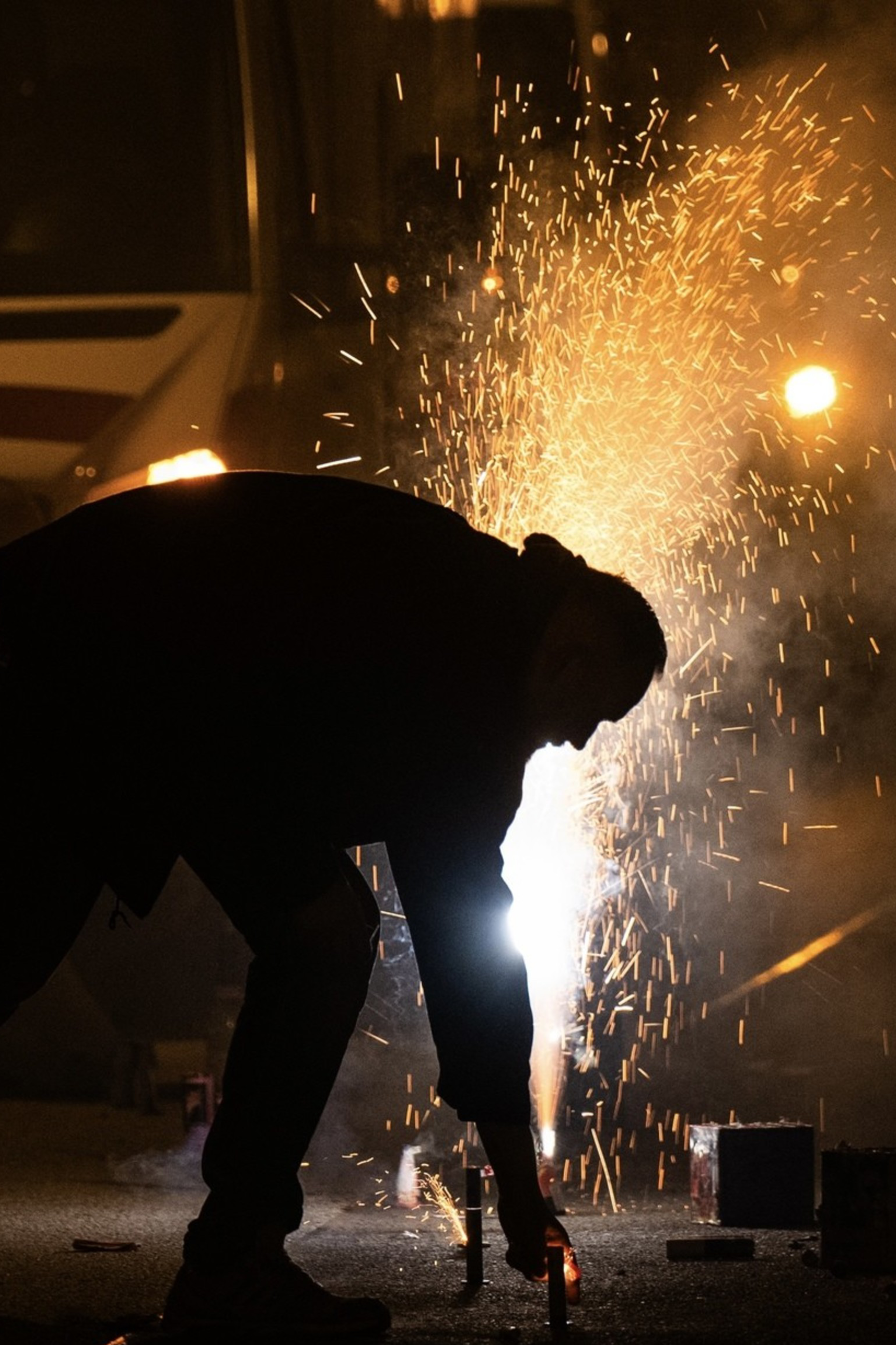 A person bends down to light fireworks in the street at night, with sparks flying and smoke filling the air, illuminating the otherwise dark surroundings.