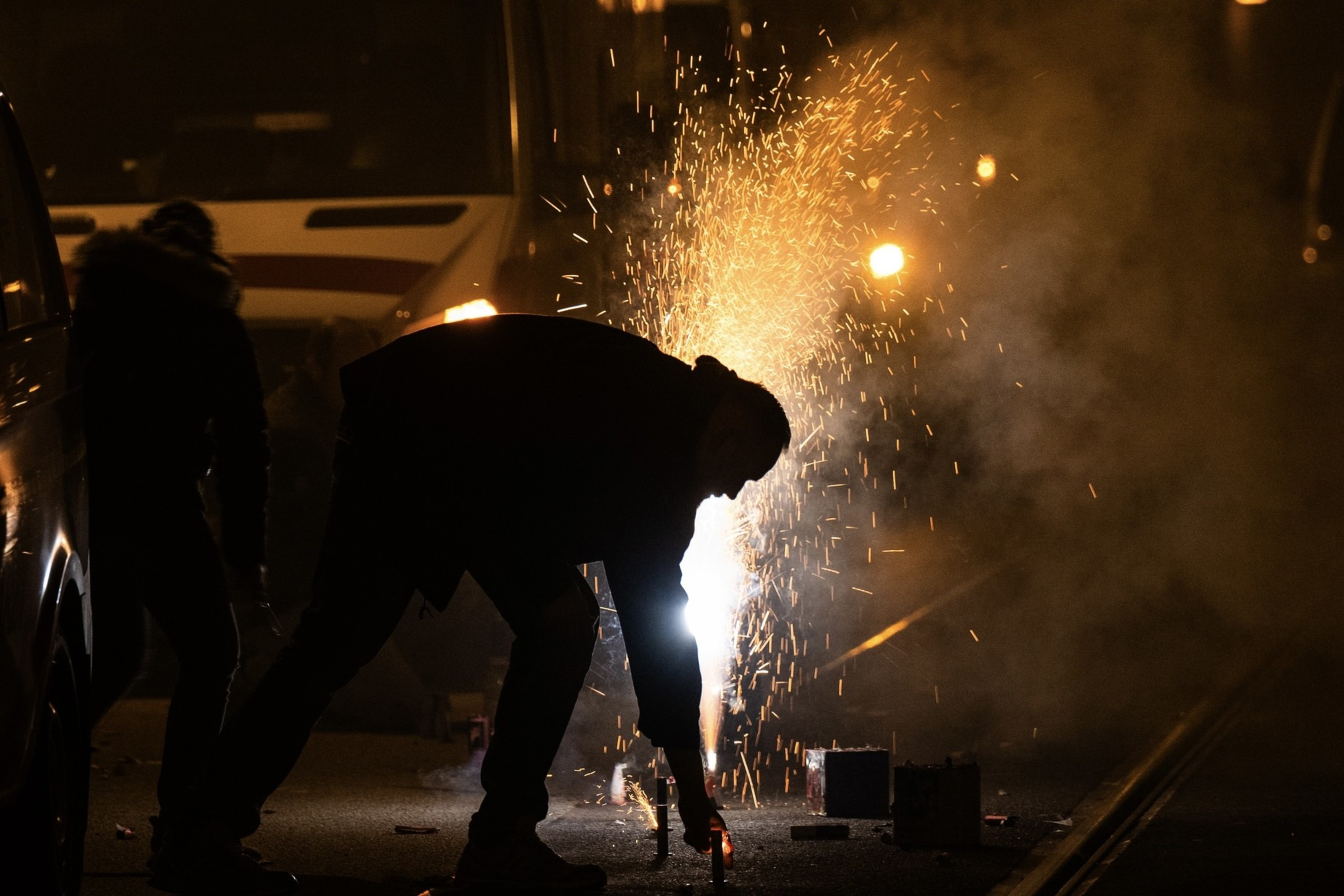 A person bends down to light fireworks in the street at night, with sparks flying and smoke filling the air, illuminating the otherwise dark surroundings.