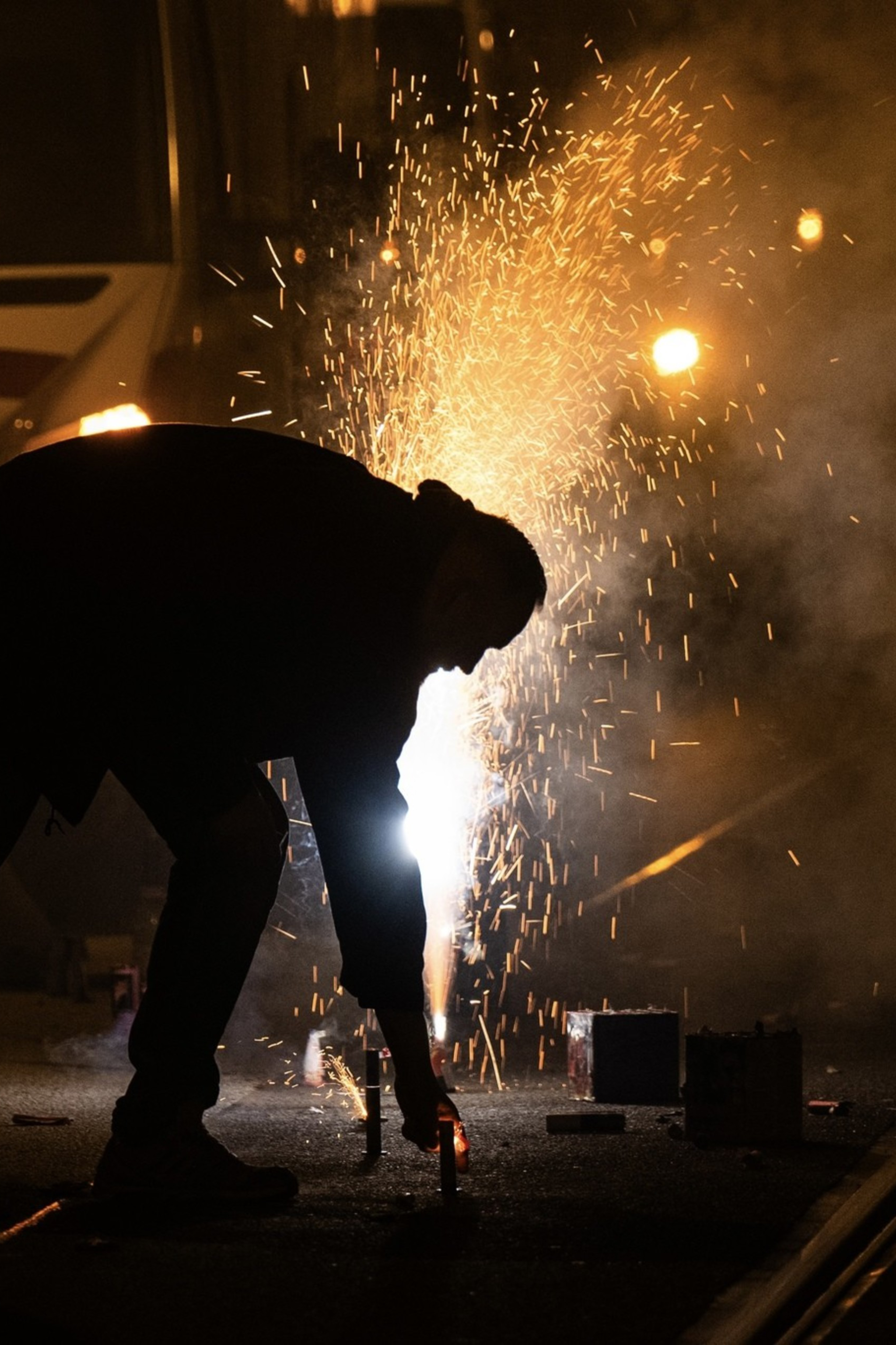 A person bends down to light fireworks in the street at night, with sparks flying and smoke filling the air, illuminating the otherwise dark surroundings.