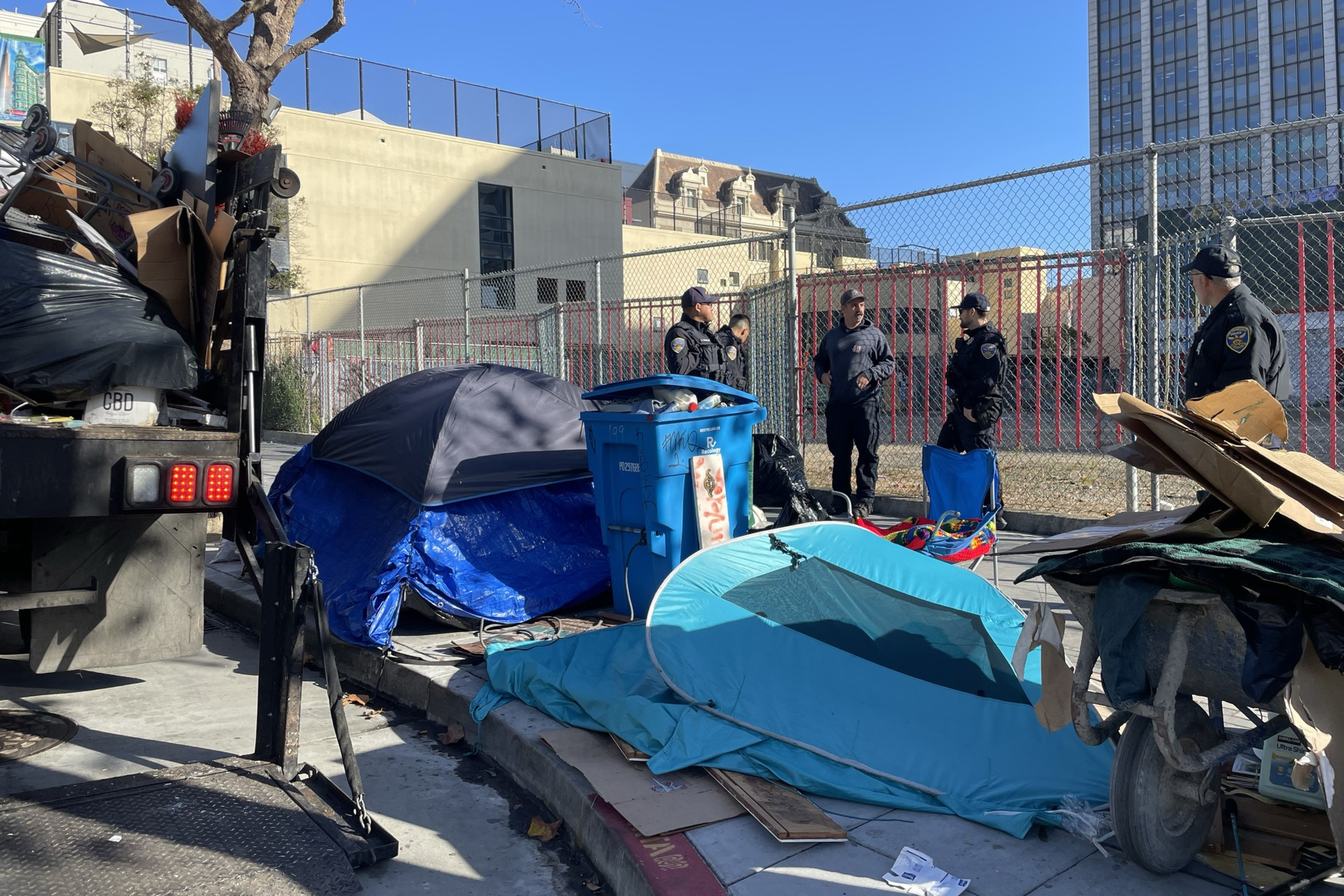 3 San Francisco Police Department (SFPD) officers stand next to a tent and small homeless encampment on the sidewalk on a sunny day.
