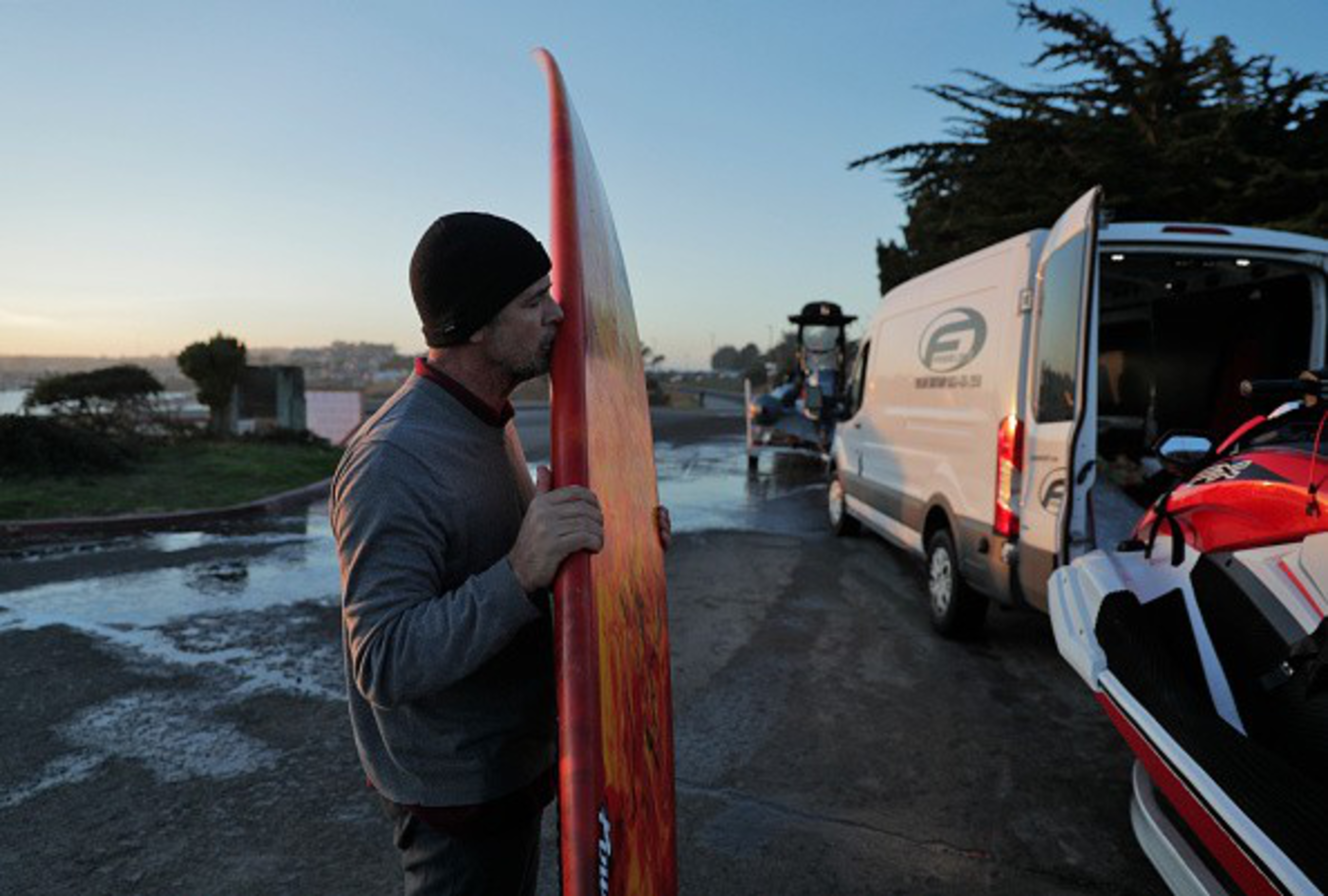 A man wearing a black beanie and gray sweater kisses a red surfboard near a white van and a jet ski at dusk by a wet road.