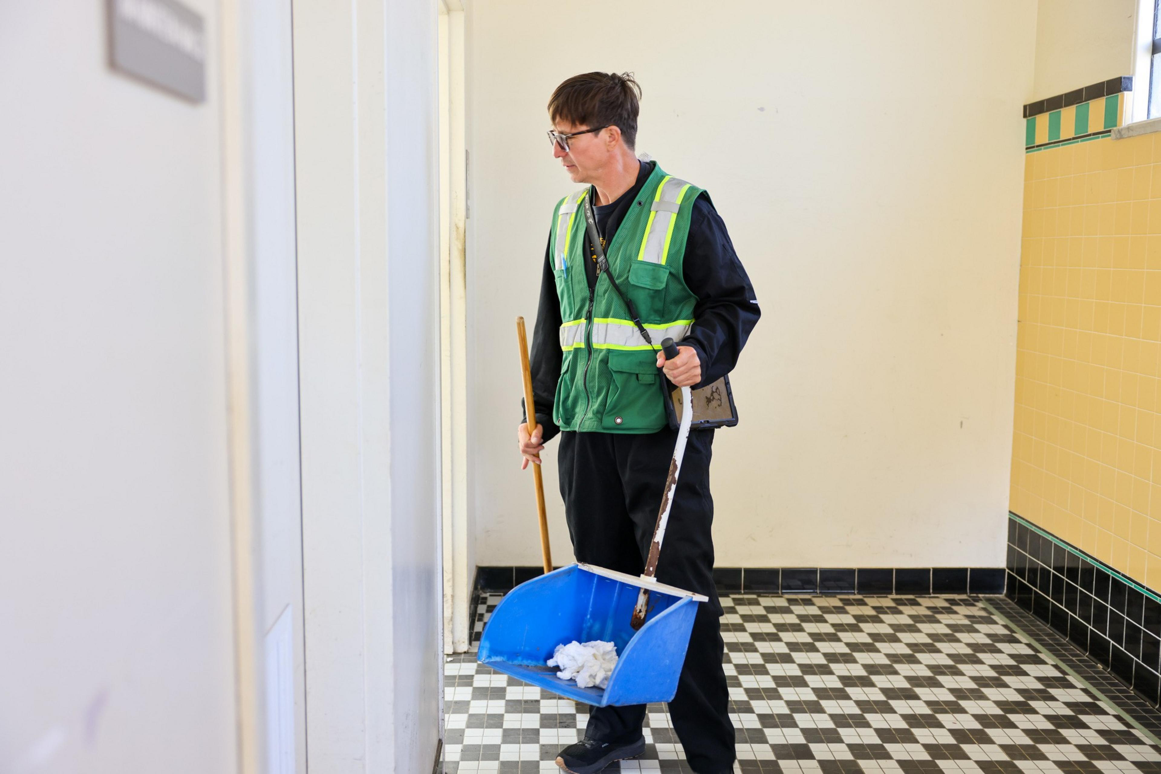 A person in a green reflective vest is holding a broom and a blue dustpan in a tiled room with yellow and white walls.