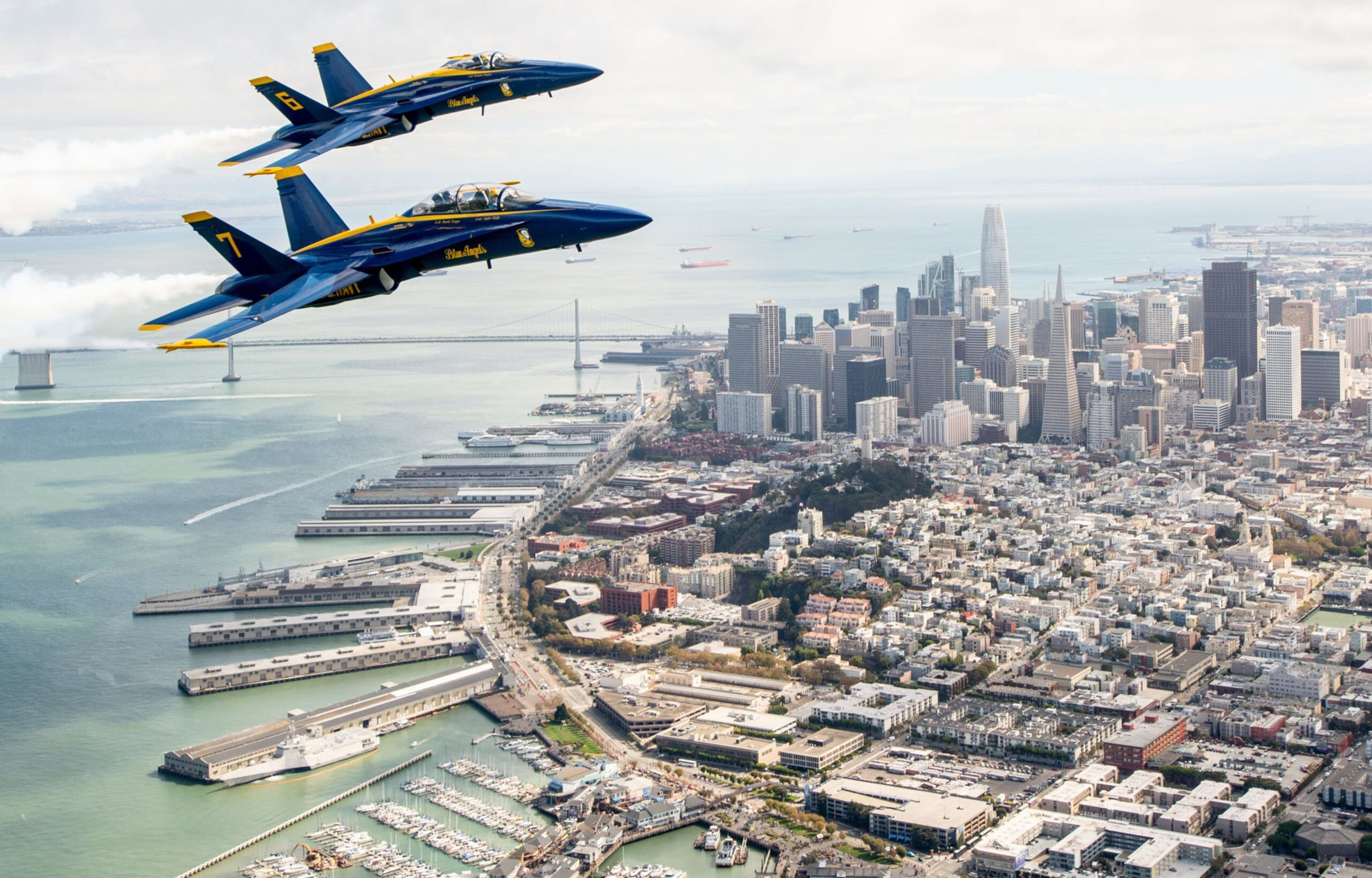 Two Blue Angels jets fly in formation above San Francisco's waterfront with the city skyline and Bay Bridge in the background.