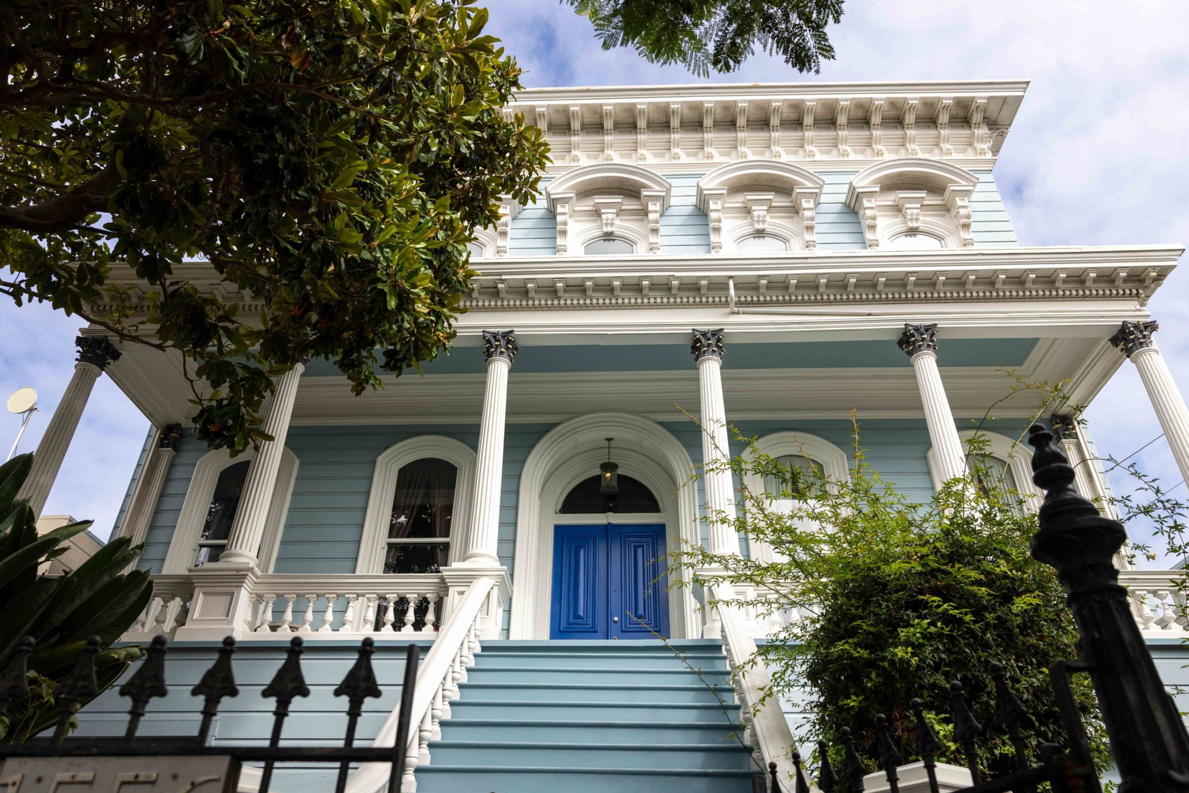 A blue Victorian house with white trim features tall white columns, a blue front door, a staircase, and lush green trees partially framing the view.