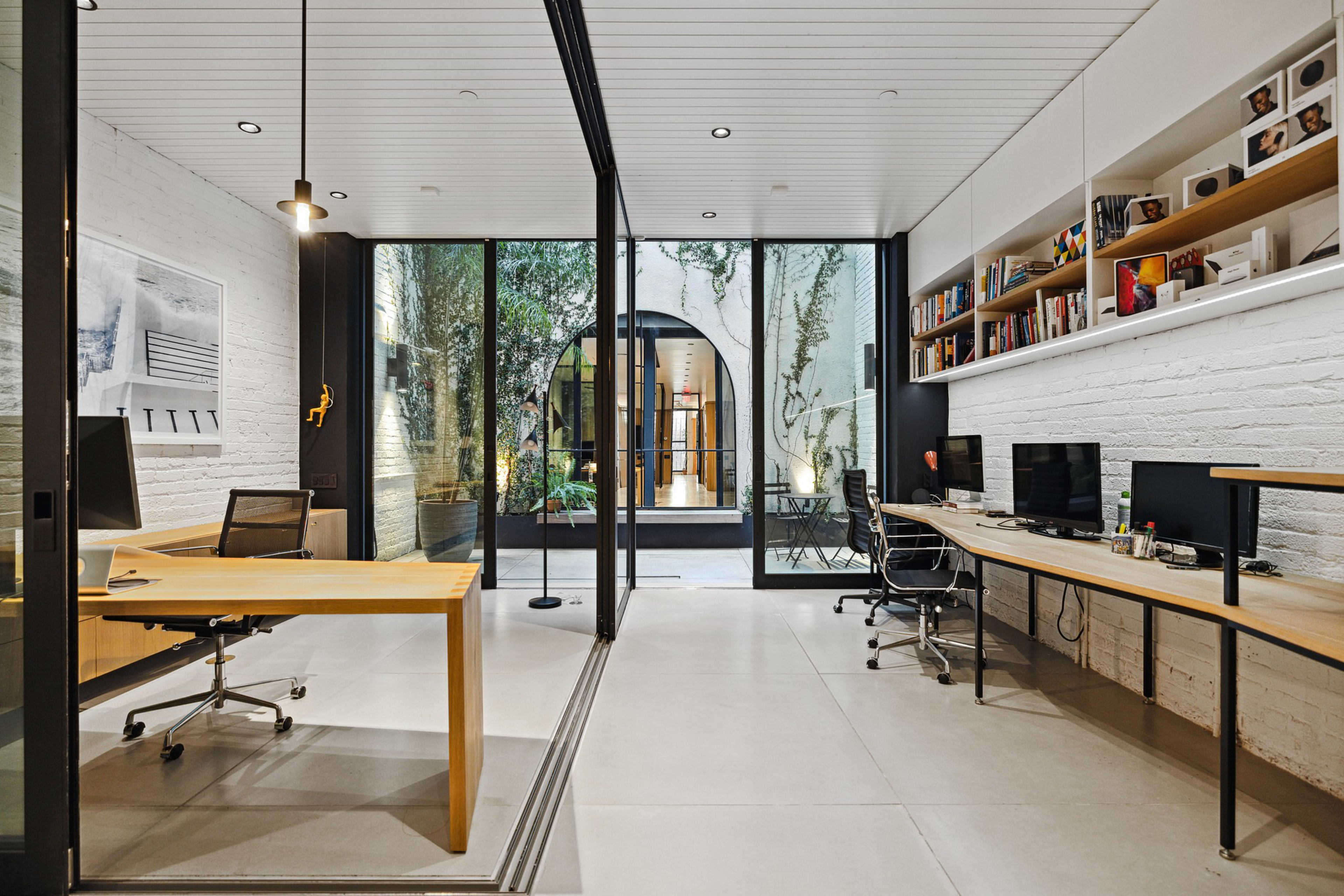 A modern office space with glass walls separates two work areas, featuring wooden desks, office chairs, computers, bookshelves, and a courtyard with greenery visible through large windows.