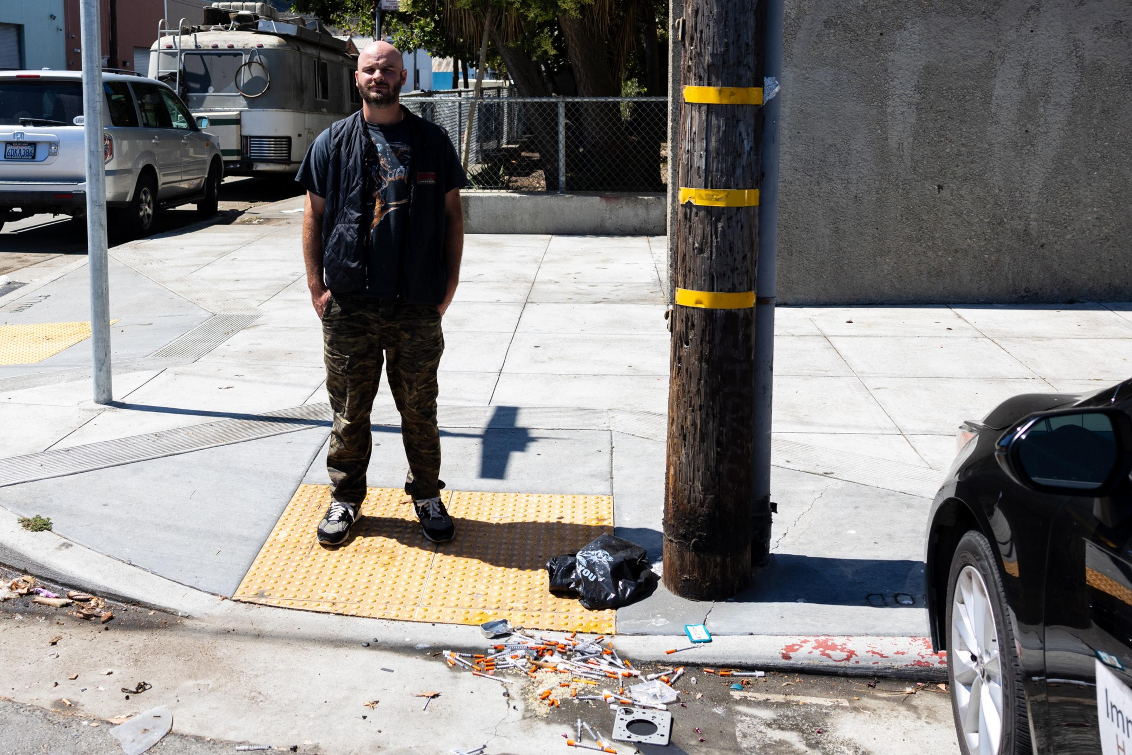 A man stands on a sidewalk near a utility pole with yellow bands, surrounded by scattered discarded syringes and a black plastic bag.