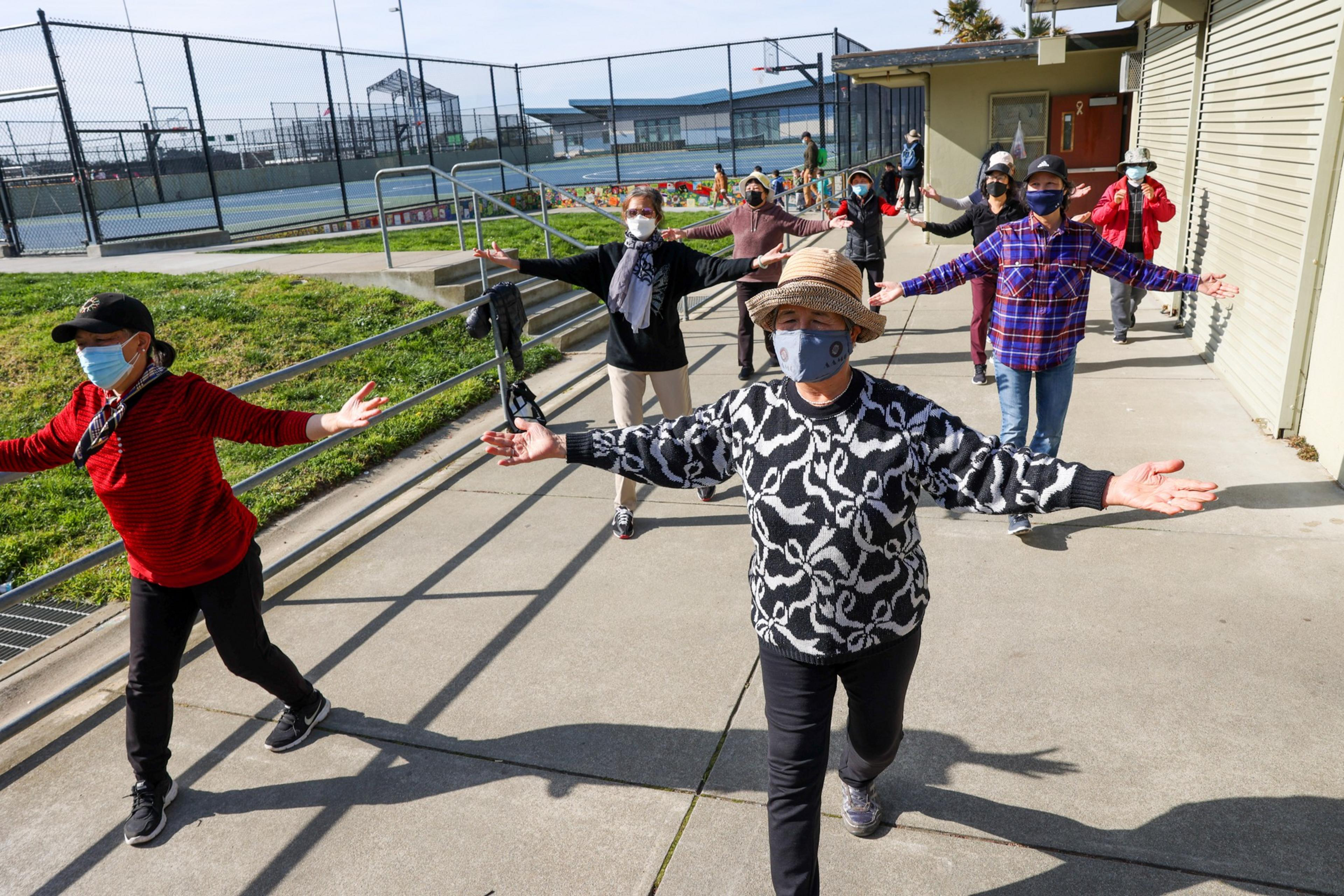 A group of people wearing masks and casual clothes stands outdoors, spaced apart with arms extended, practicing a balance or stretching exercise.