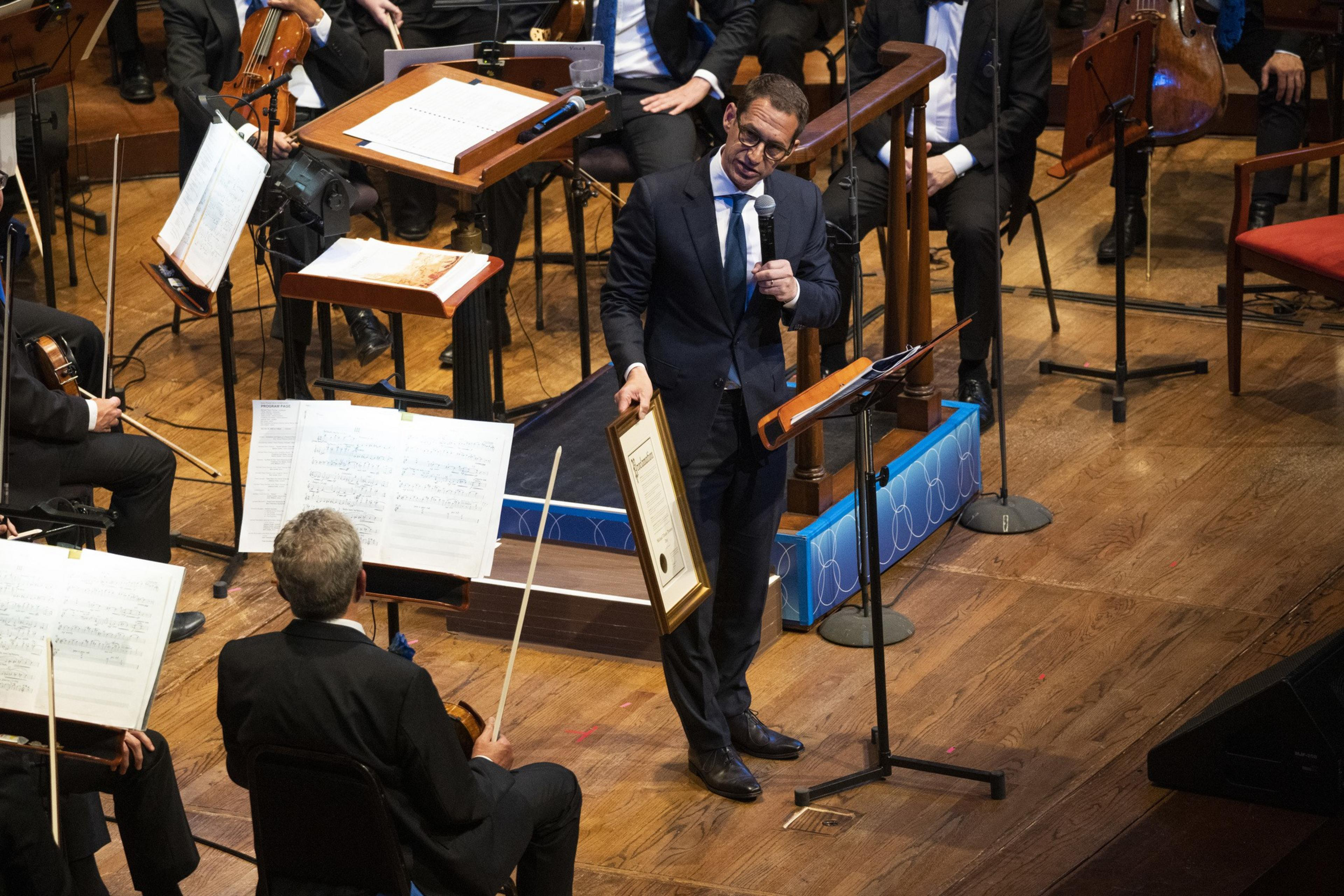 A man in a suit is speaking into a microphone on stage, holding a framed certificate. Musicians with sheet music are seated around him.
