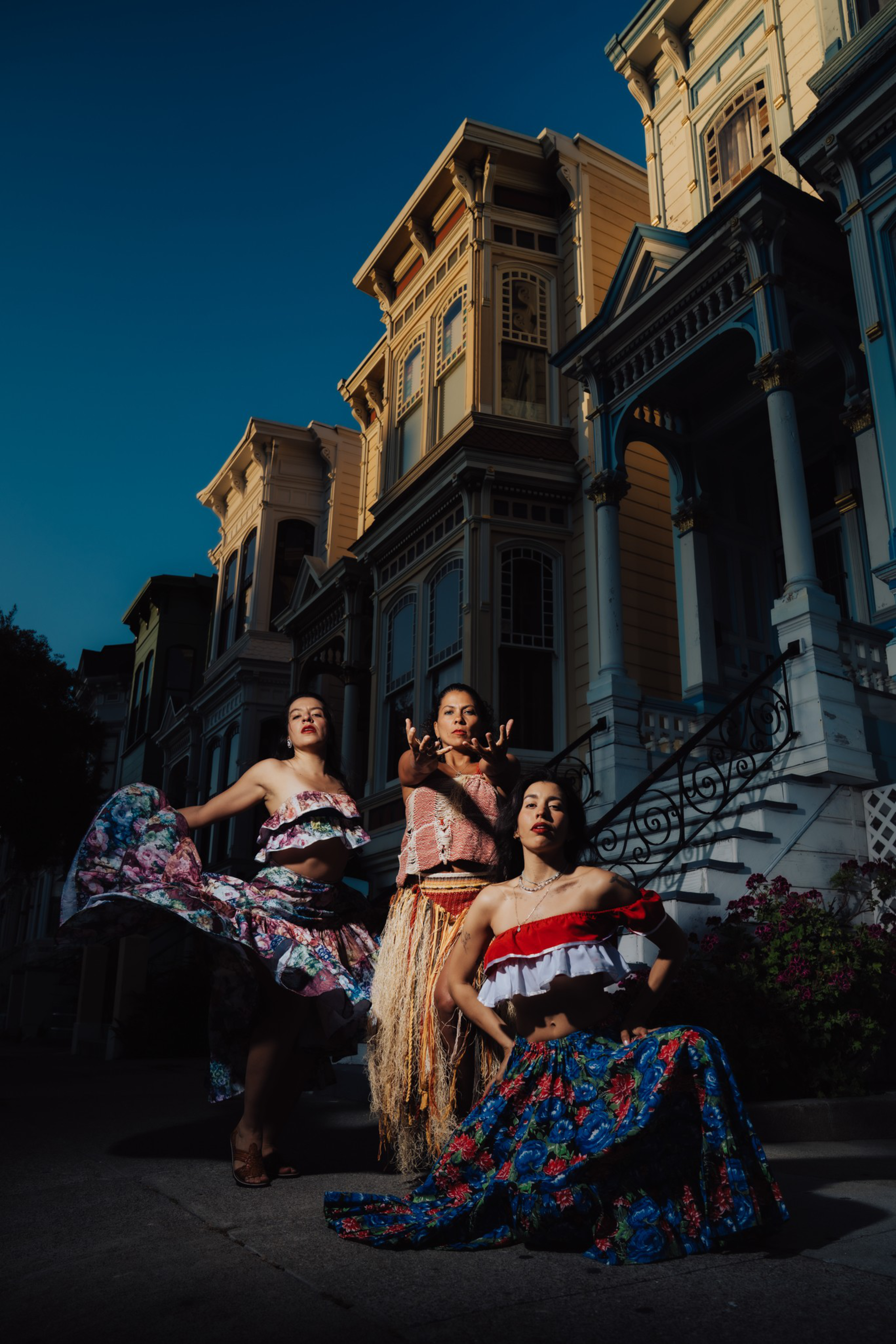 Three women pose confidently in colorful, traditional dresses on a street with tall, ornate Victorian houses in the background at dusk.