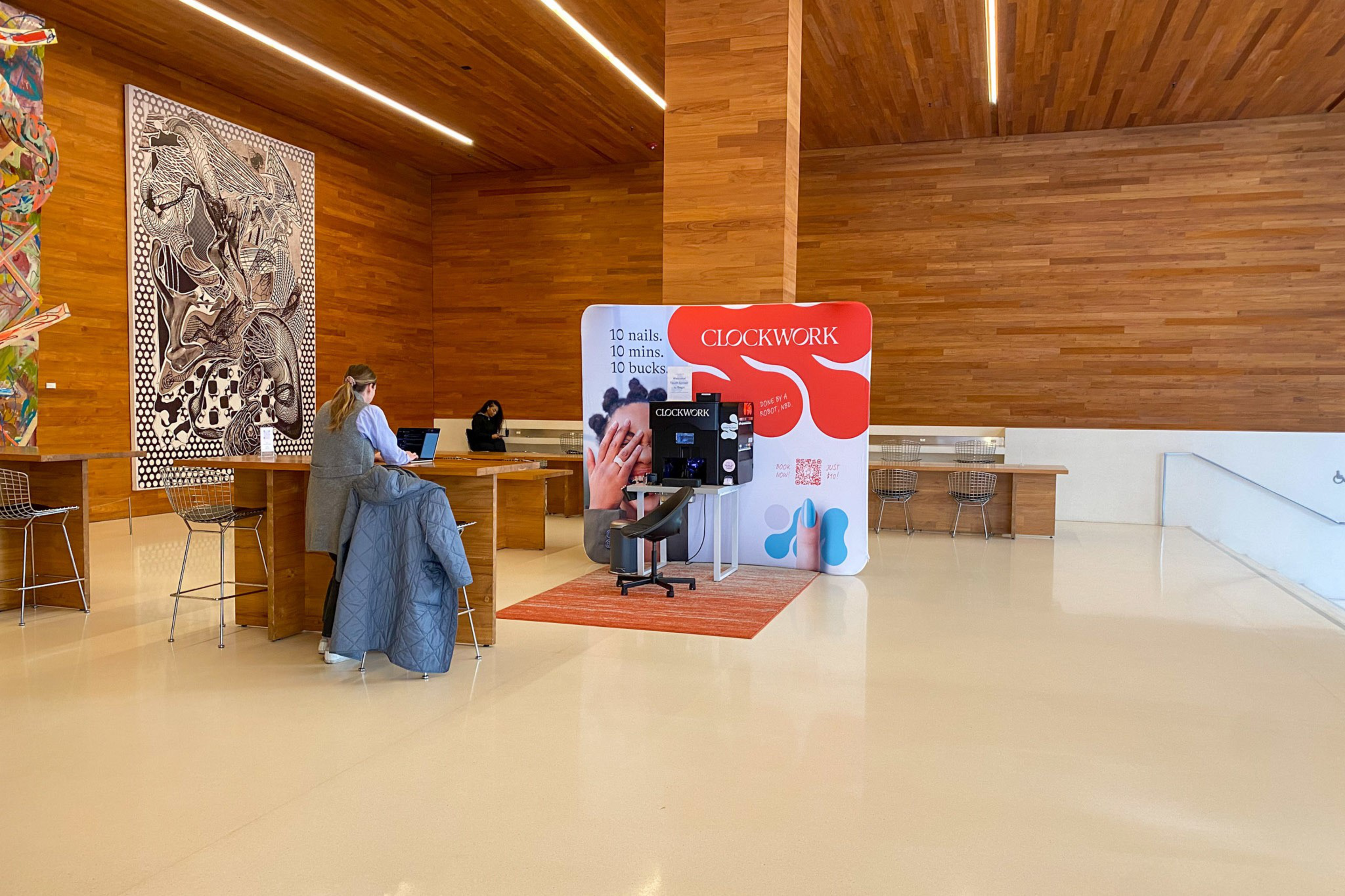 The image shows a modern space with wooden walls, featuring Clockwork's nail service booth. A person is using a laptop at a wooden table, and art pieces adorn the walls.