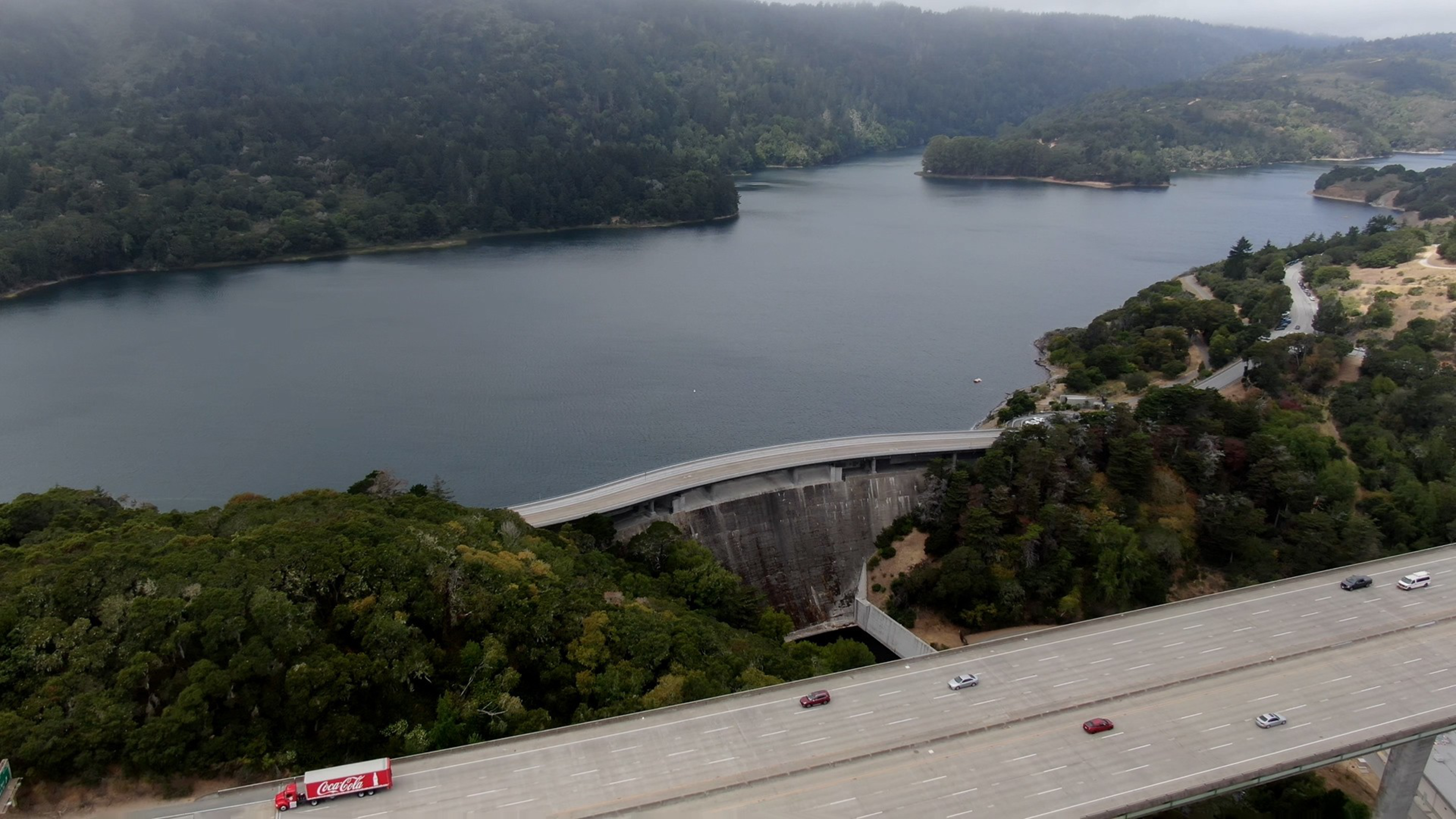 A large dam holds back a calm lake surrounded by forested hills, with a highway bridge and several vehicles crossing in the foreground.