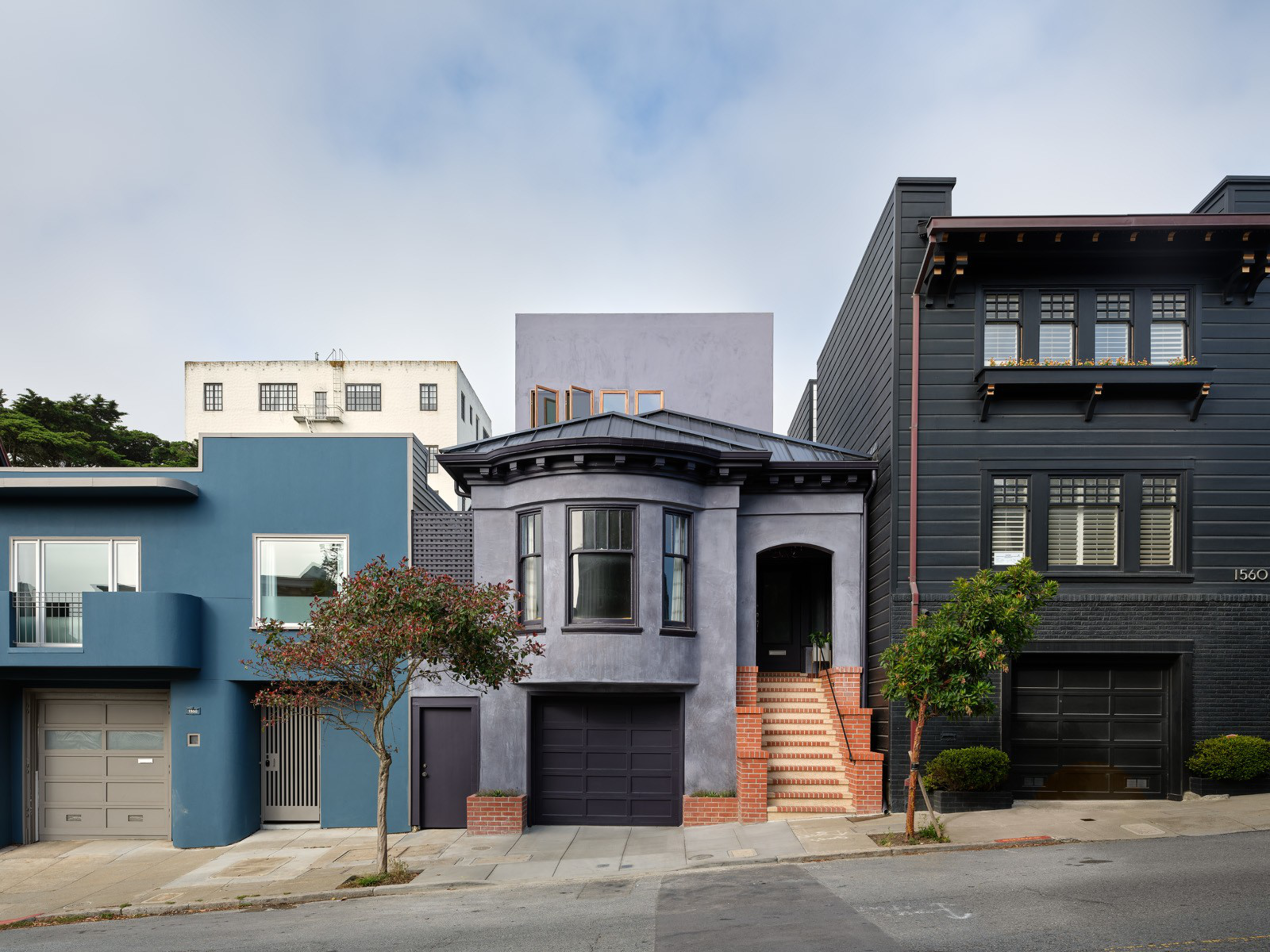 Three adjacent houses with garages and front trees stand on a sloped street under a partly cloudy sky.