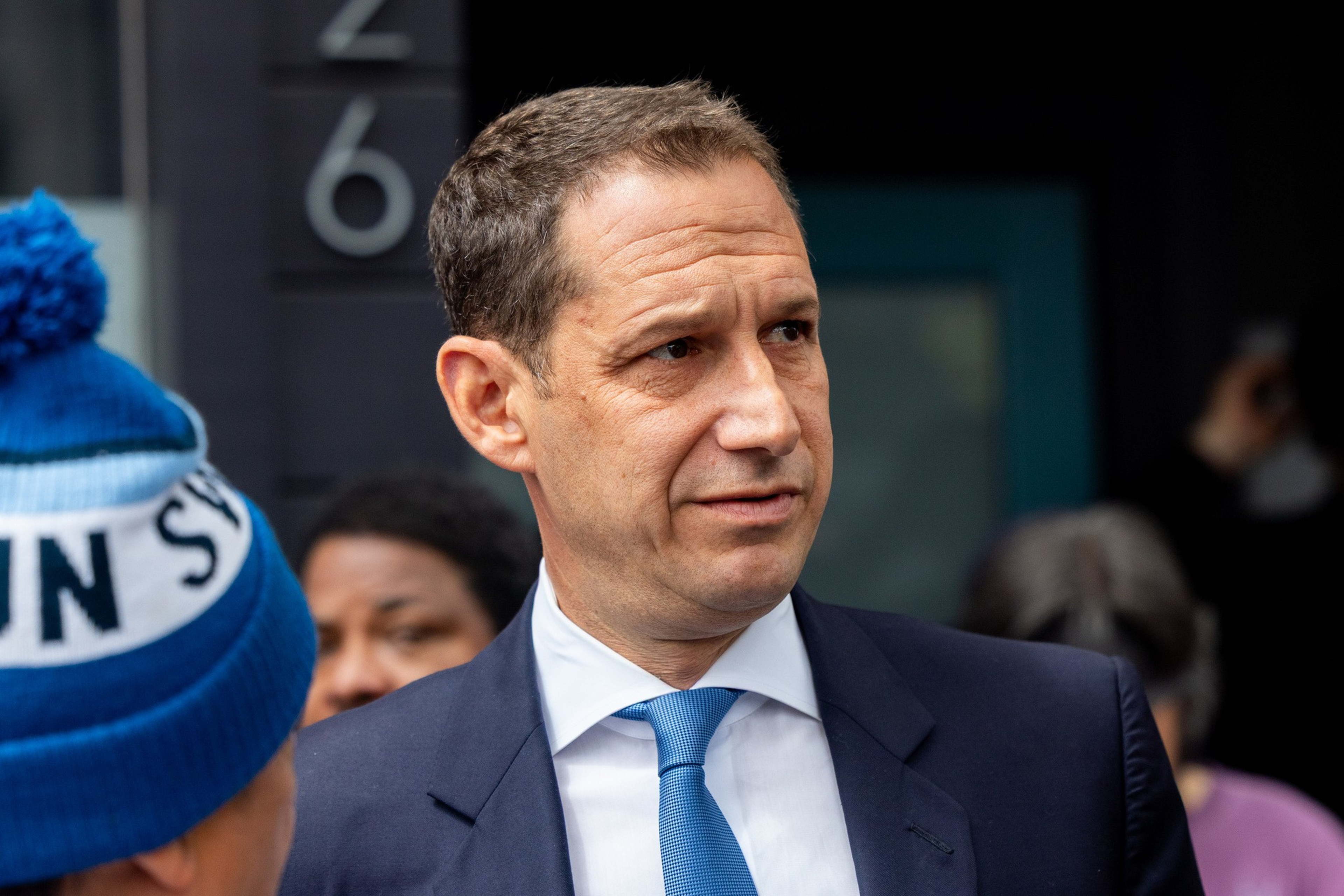A man in a blue suit and blue tie looks to his right, with blurred people and a building number 26 in the background.