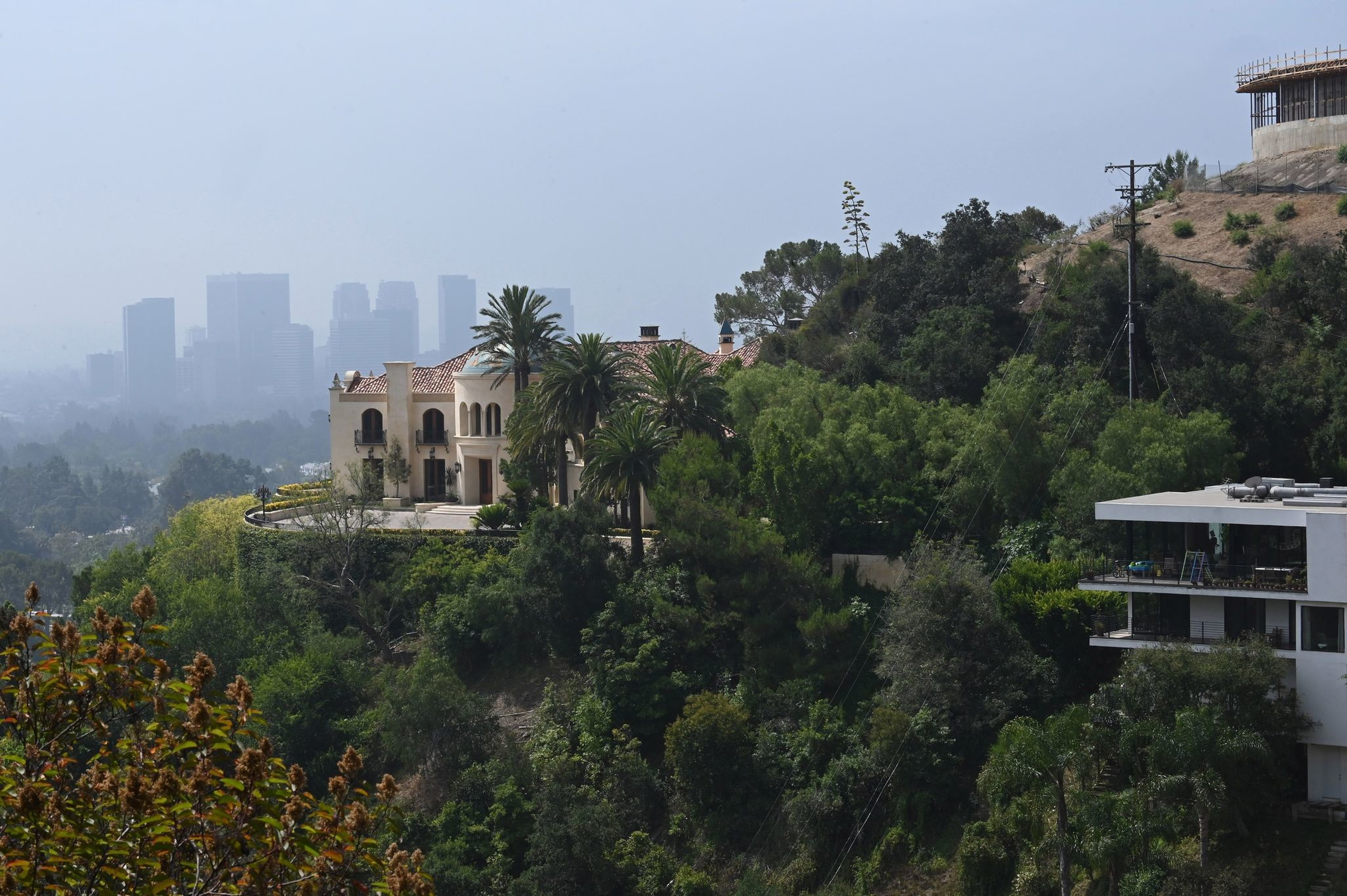 A large Mediterranean-style house sits on a hillside surrounded by palm trees and dense greenery, with city skyscrapers visible in the hazy background.