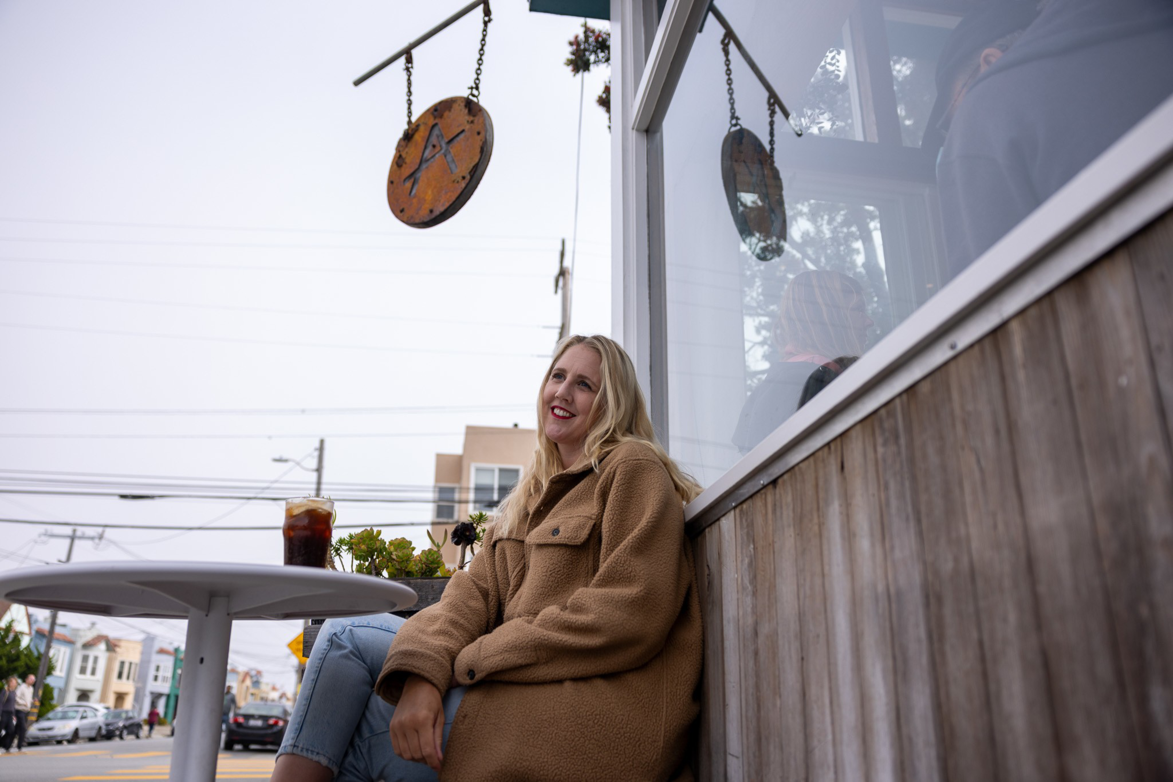 A woman in a tan coat sits outside a cafÃ©, smiling. A glass of dark iced beverage is on the table, and a round cafÃ© sign with an “A” hangs above her.