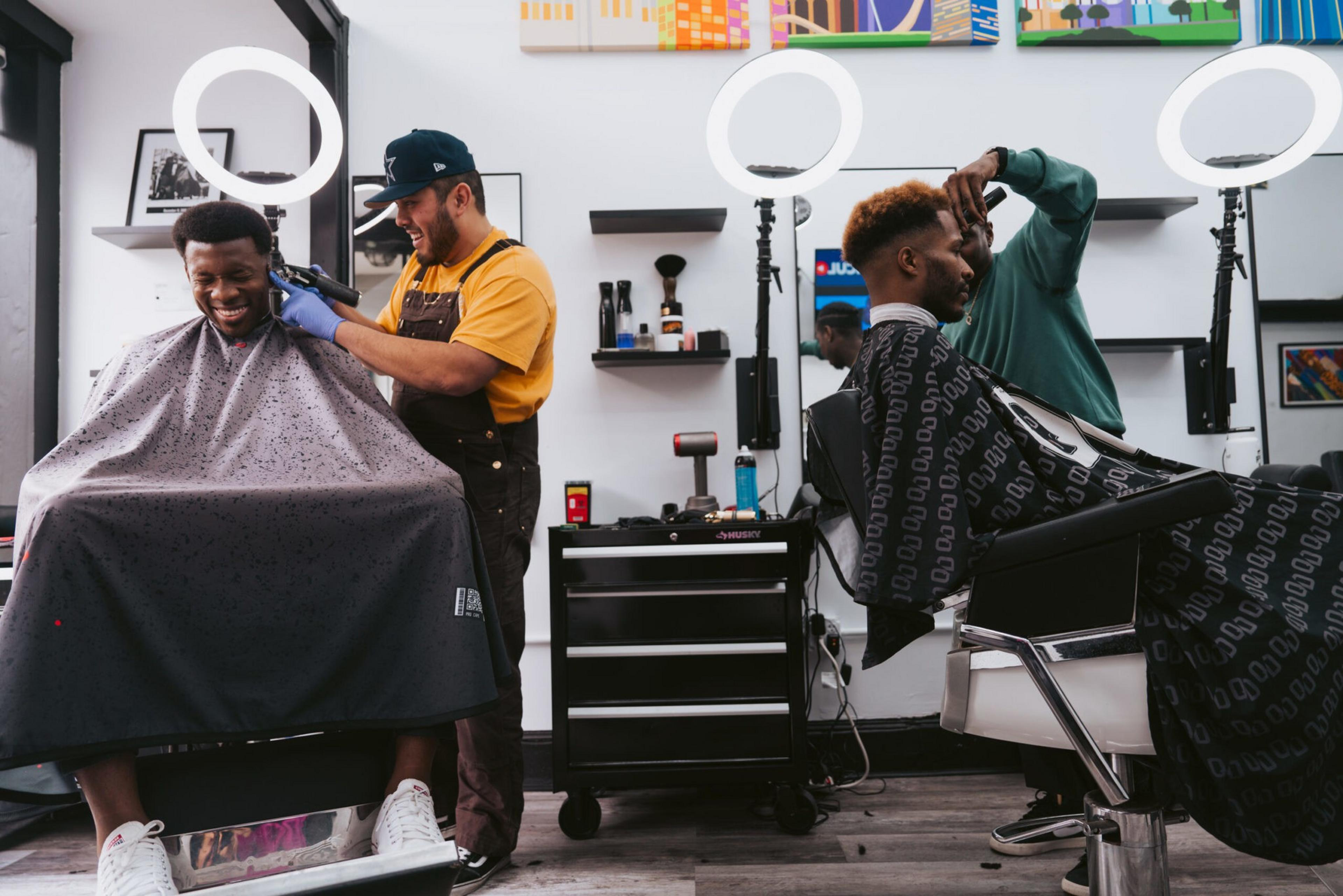 Two men get haircuts from barbers in a modern shop with ring lights, shelves, and colorful wall art in the background.