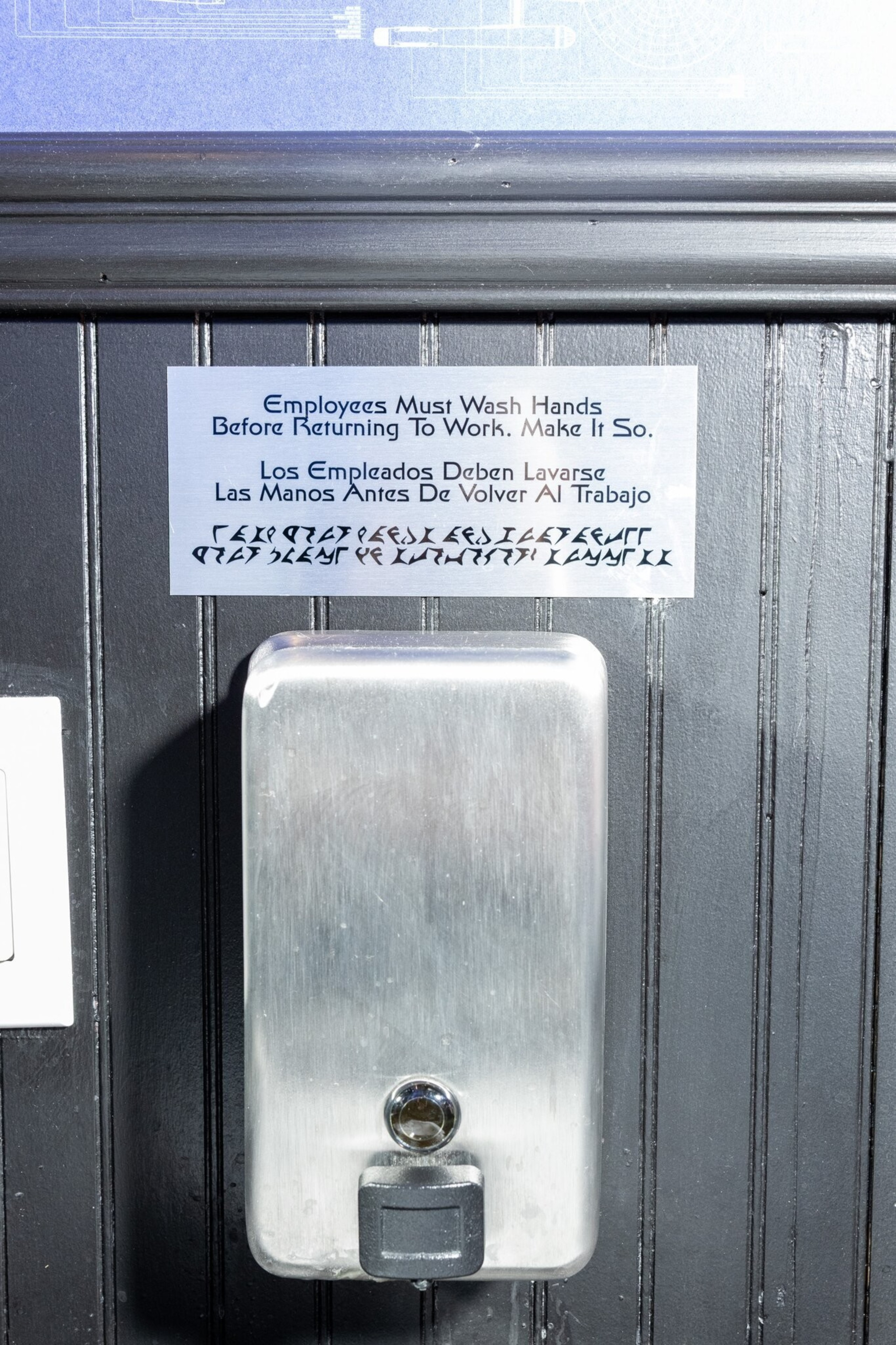 A stainless steel soap dispenser is mounted on a wall under a sign instructing employees to wash hands before returning to work, written in English and Spanish.