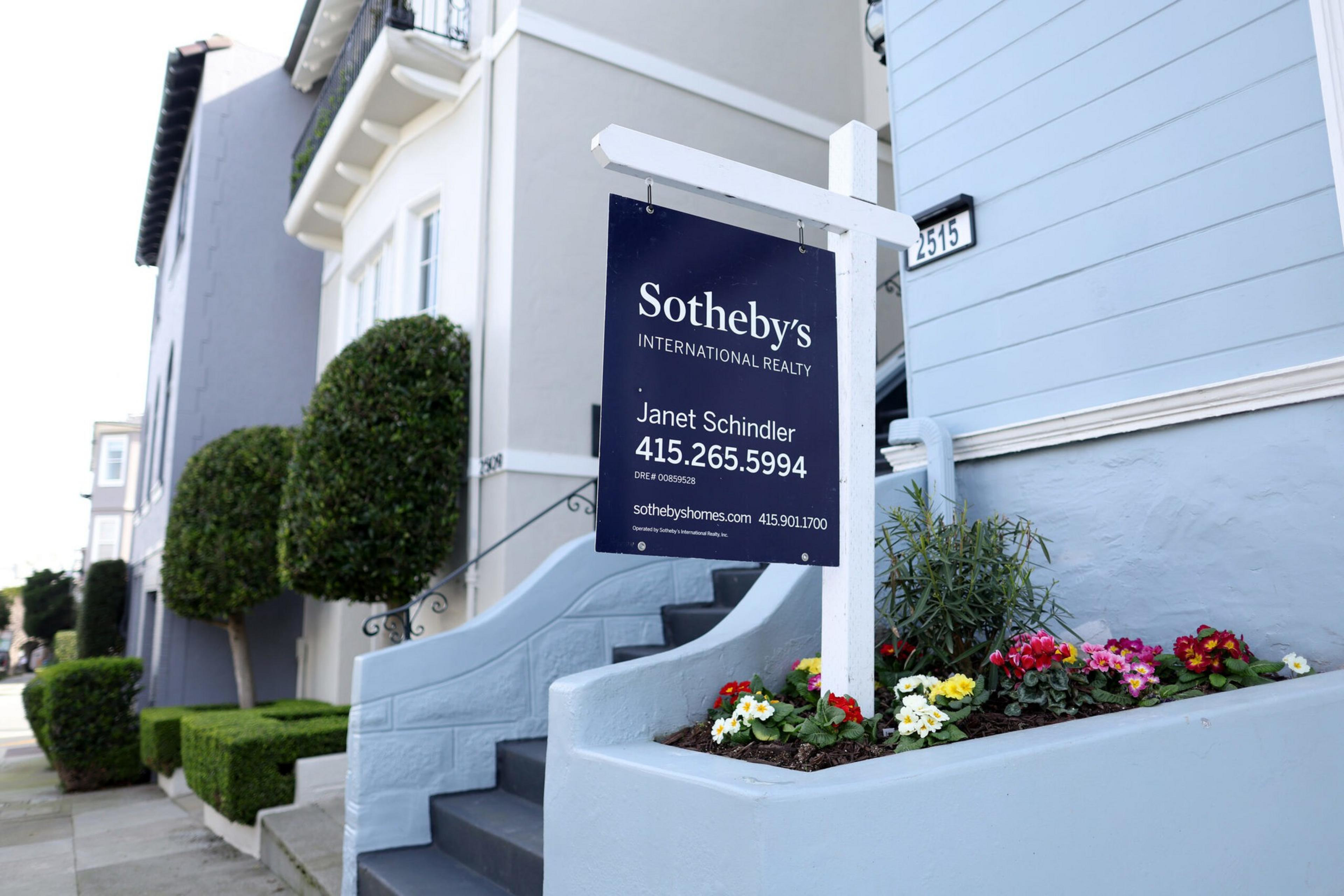 A Sotheby's Realty sign is placed in front of a light blue house with steps and colorful flowers in a small garden bed nearby.
