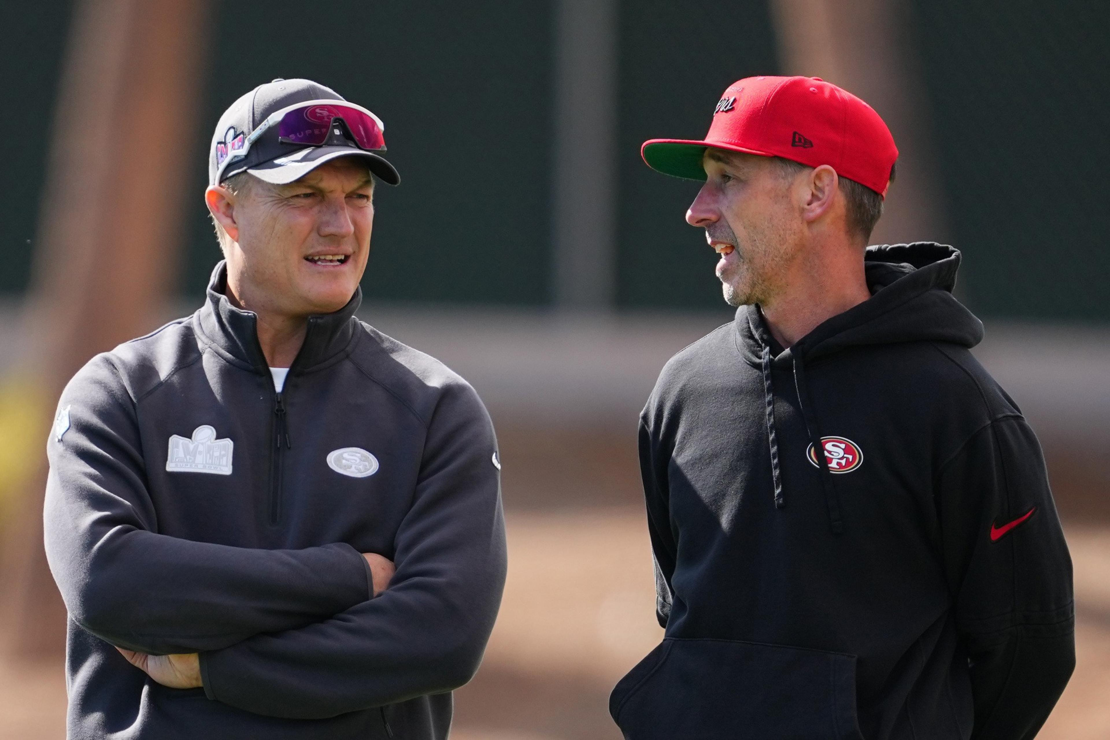 Two men are talking outdoors, wearing San Francisco 49ers gear. One has a gray jacket and sunglasses on his cap, while the other wears a red cap and black hoodie.