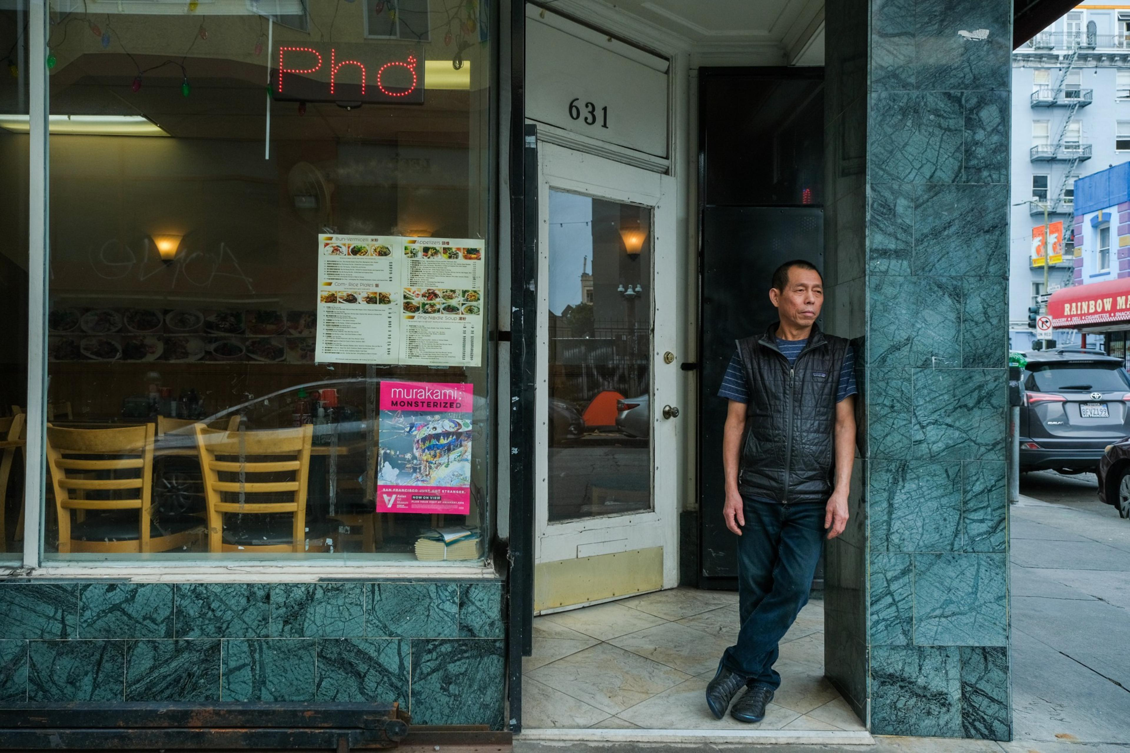 A man stands by an open door of a Pho restaurant, with a menu and a colorful sign in the window.