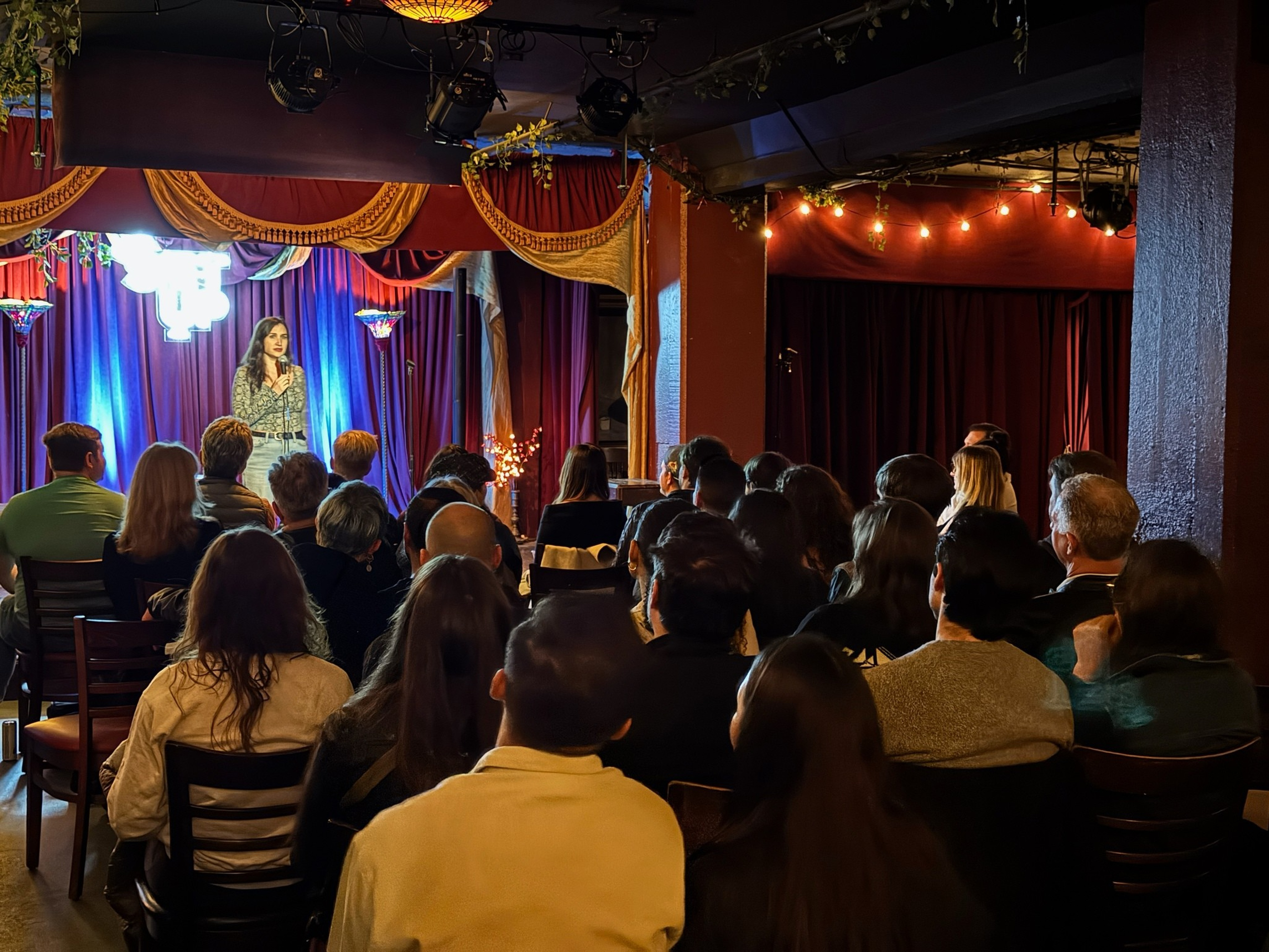 A woman stands on stage speaking into a microphone before a seated audience in a cozy, dimly lit venue with red curtains and decorative lights.