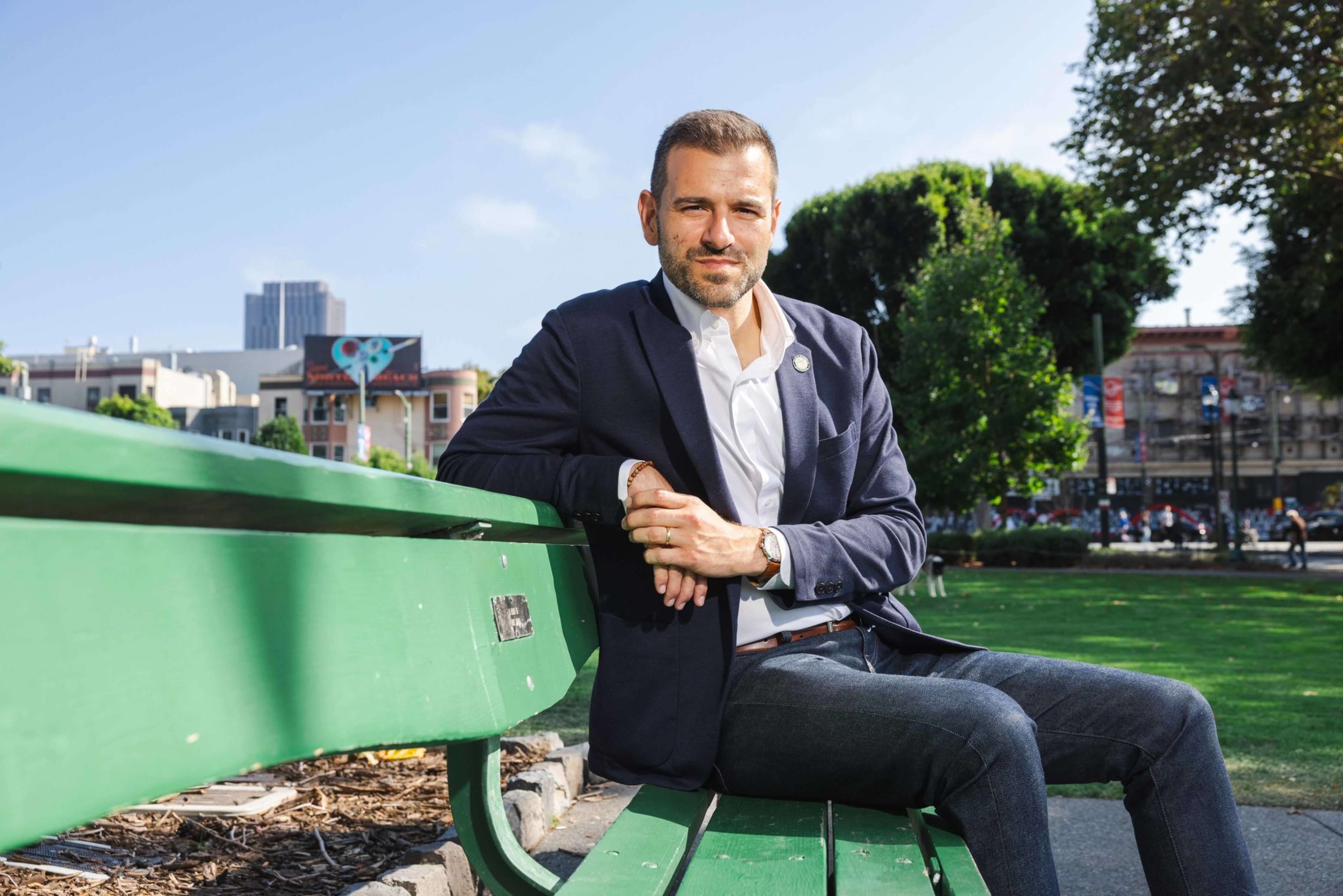 A man in a dark blazer and white shirt sits on a green park bench, with trees and urban buildings visible in the background.
