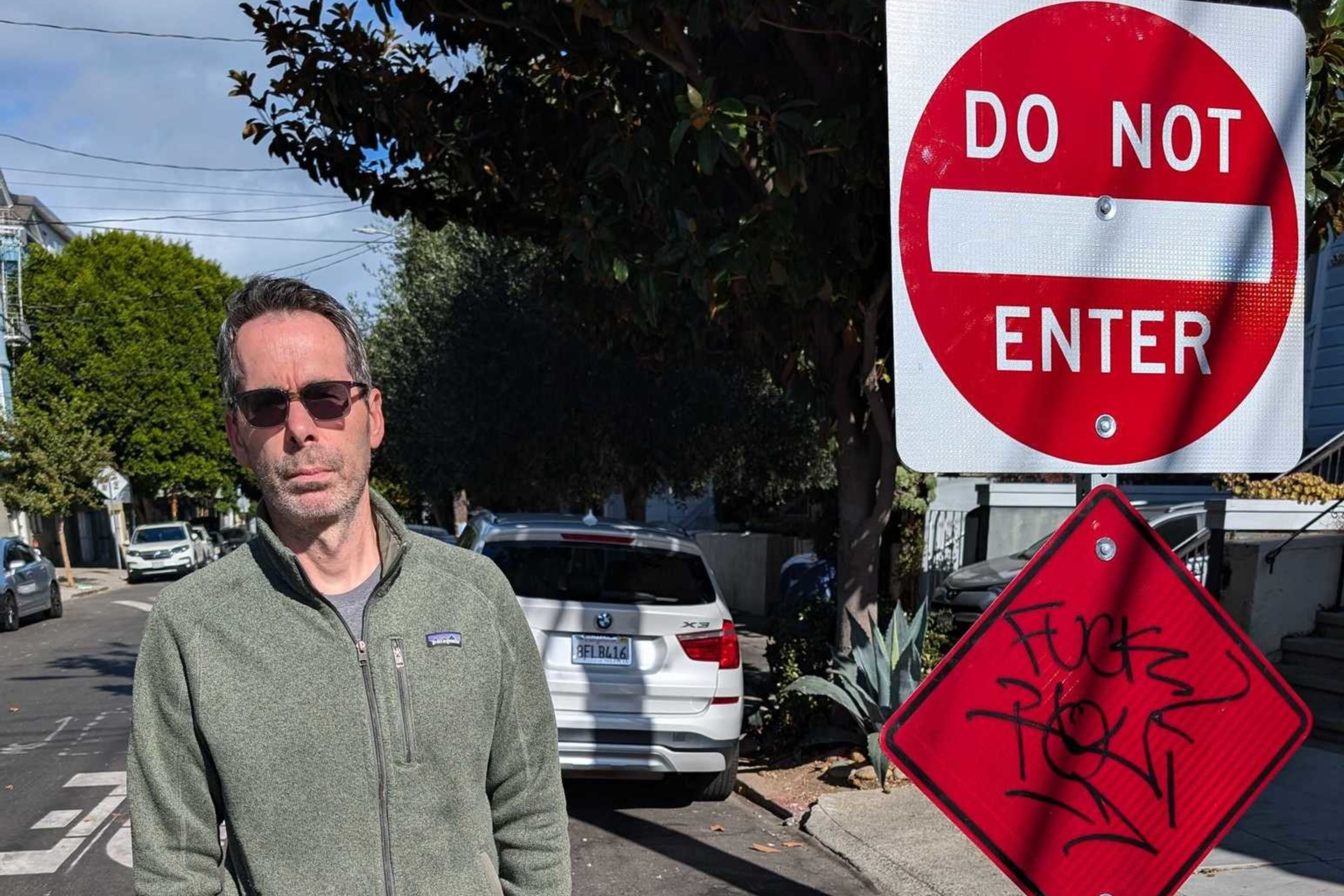 A person in sunglasses stands beside a "Do Not Enter" sign on a sunny street. They wear a green jacket and dark pants, with a car and trees in the background.