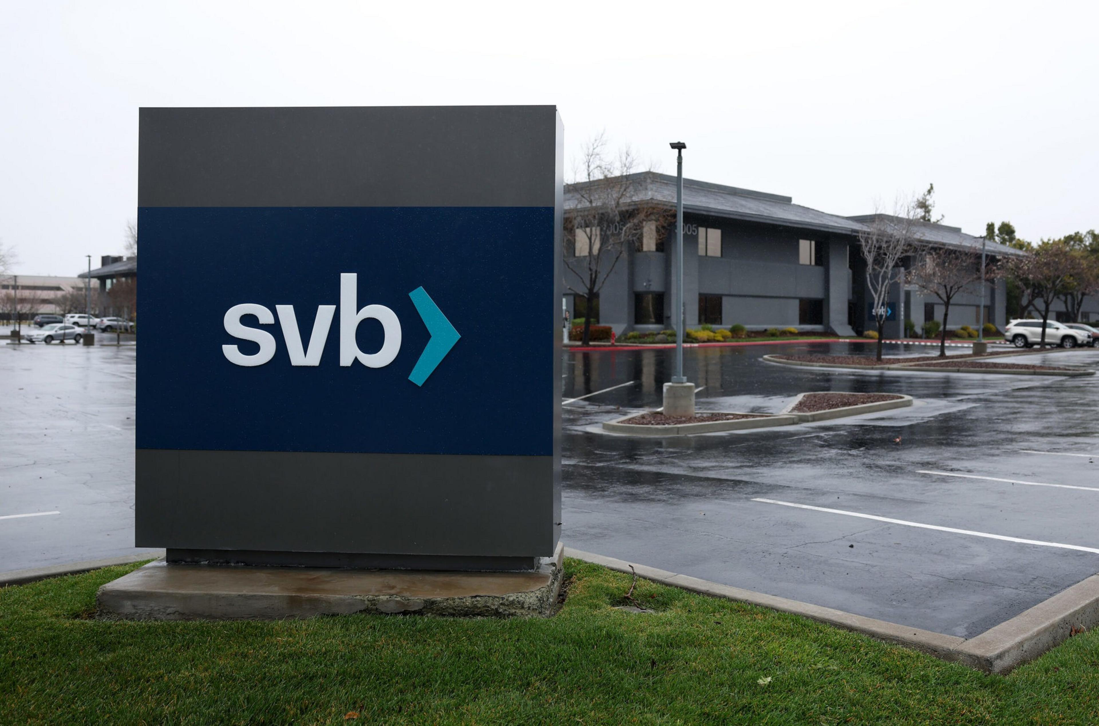 A sign with "svb" in blue sits in front of a gray office building on a rainy day, with empty parking spaces and bare trees in the background.