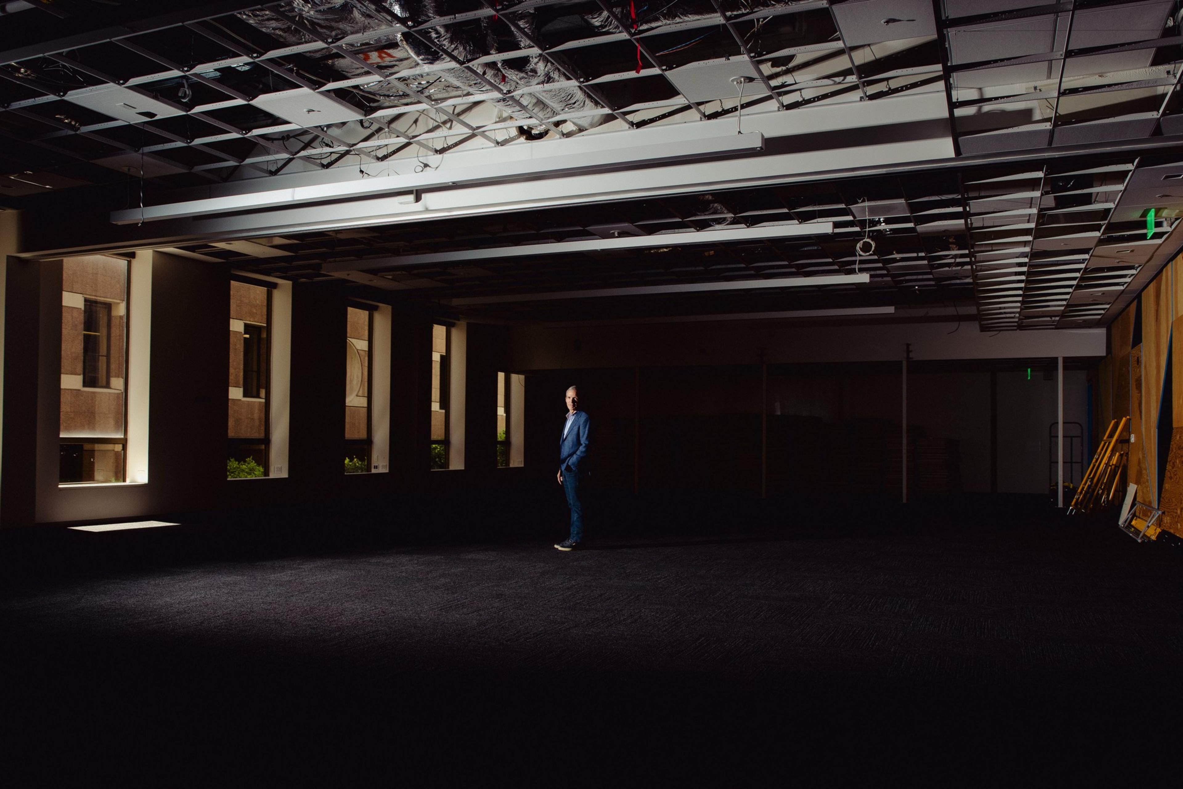 A person in a blue suit stands in an empty, dimly lit room with exposed ceiling panels. Light filters in from windows on the left, casting shadows.