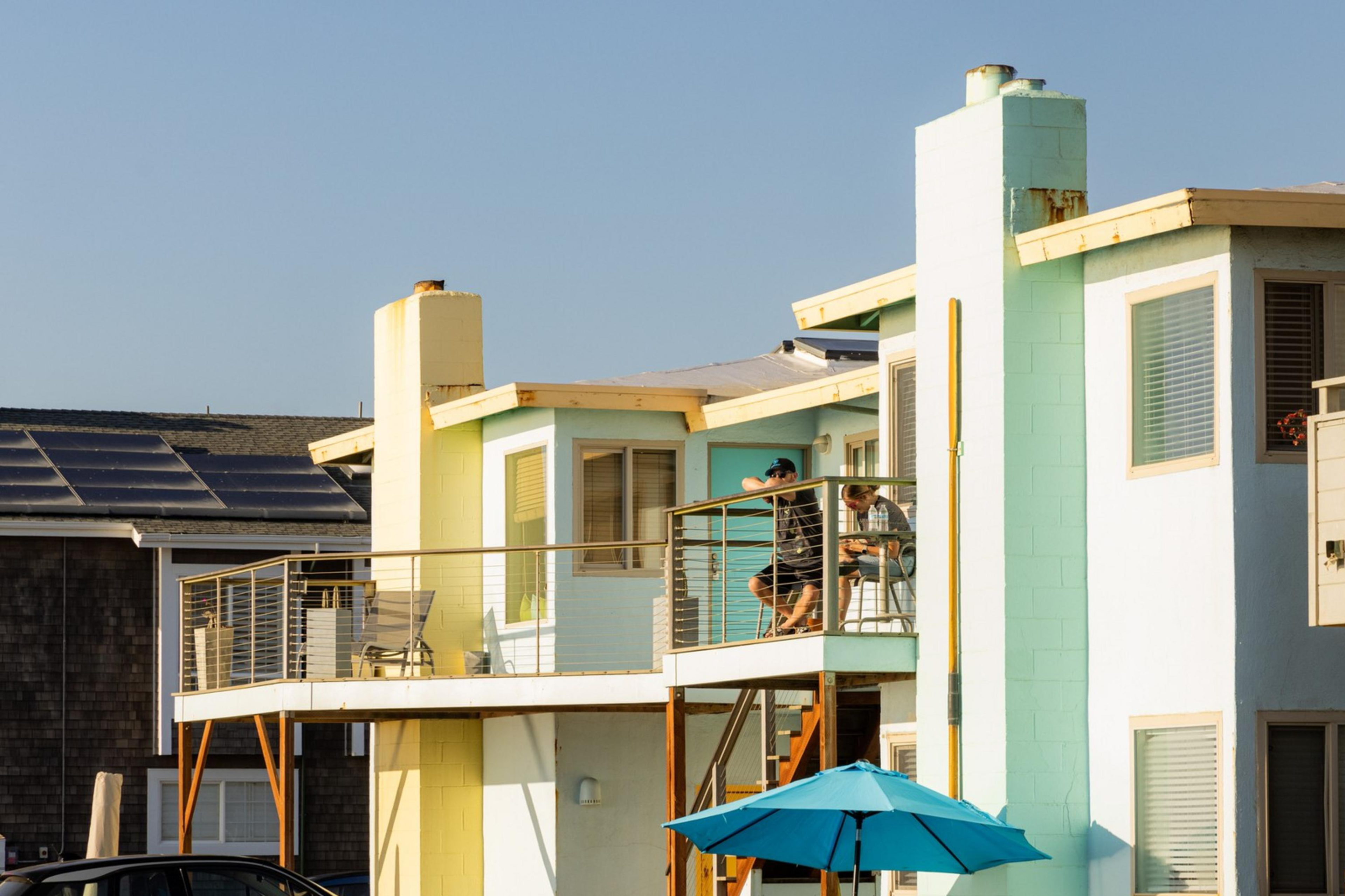 There are pastel-colored beach houses with balconies and a blue umbrella outside. People are sitting on one of the balconies, enjoying the sunny weather.