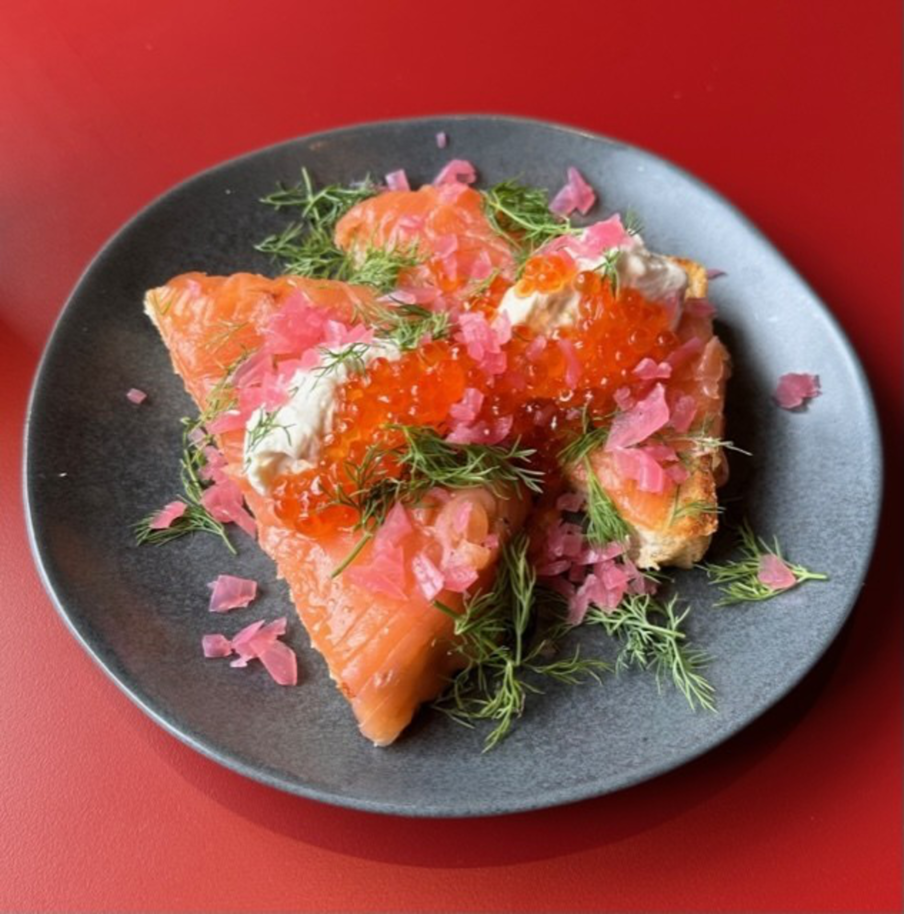 Smoked salmon on toasted bread topped with cream, red fish roe, pink pickled onions, and fresh dill on a dark plate against a red background.