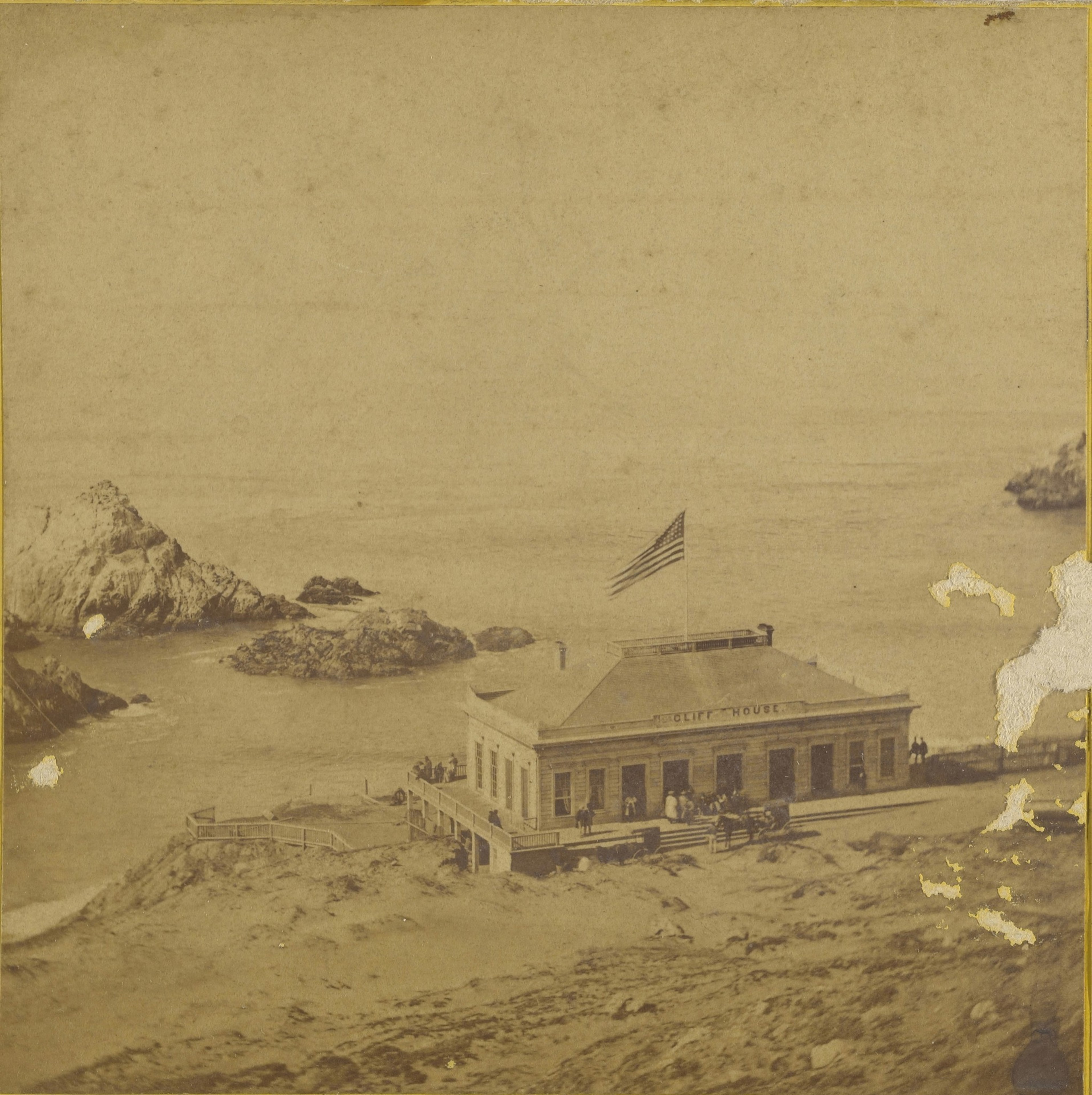 A coastal building with an American flag flying on top sits near rocky cliffs by the sea, with several people gathered around its porch.