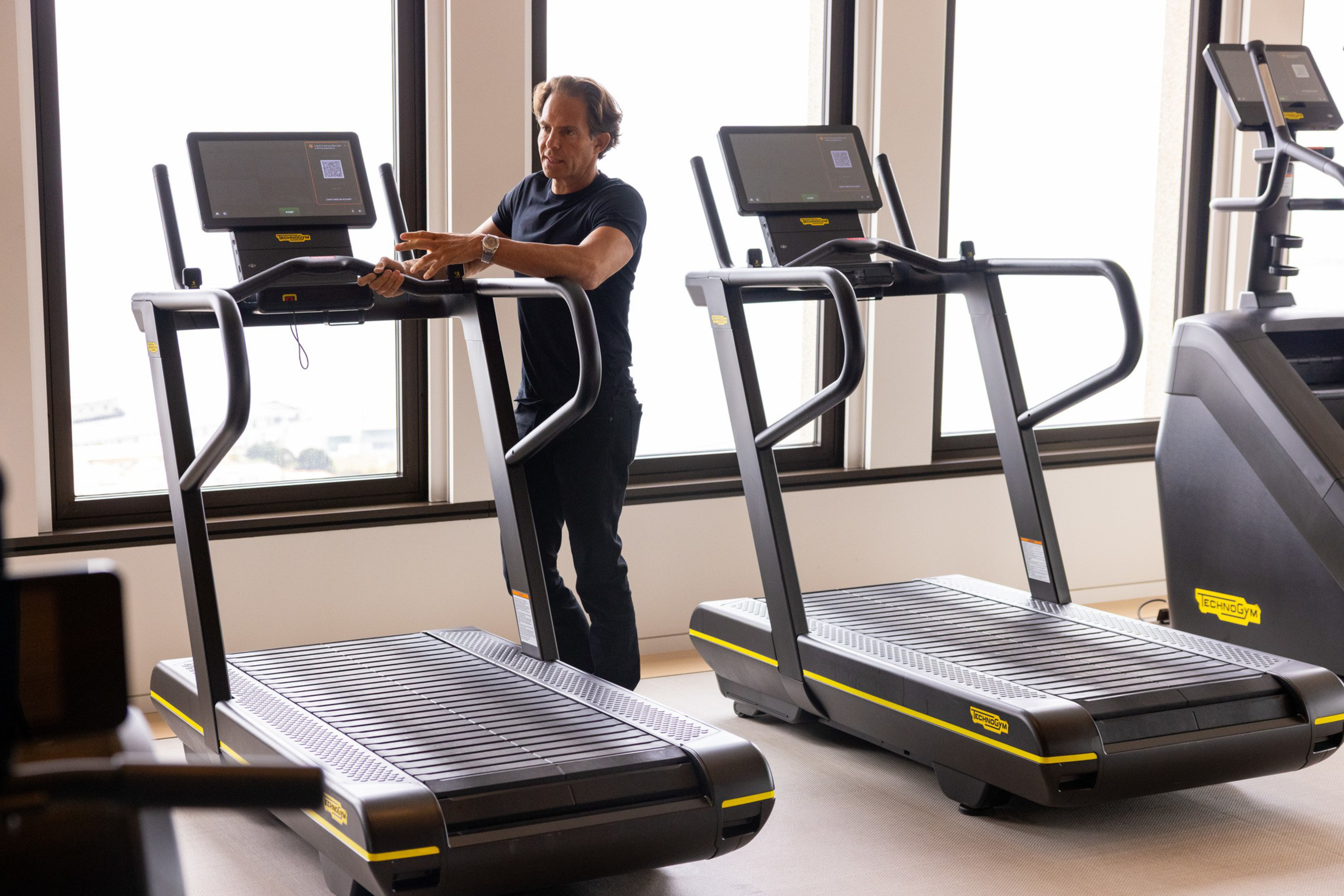 A man in a black shirt and pants stands between two treadmills, gesturing with his hands in a bright room with large windows.
