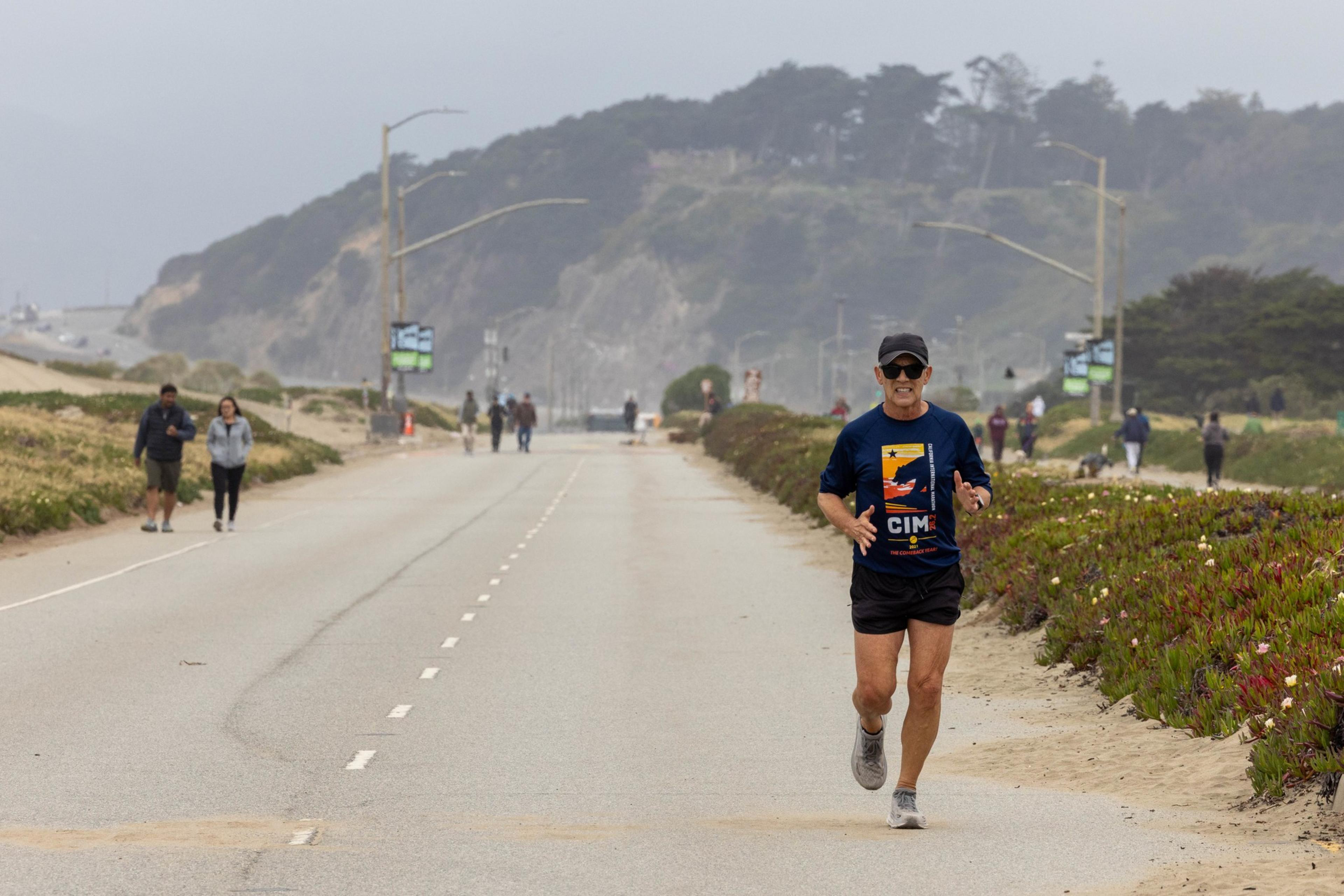 A person is jogging on an empty road near the beach, wearing a blue shirt and black cap. Other people are walking in the background, with hills visible behind them.