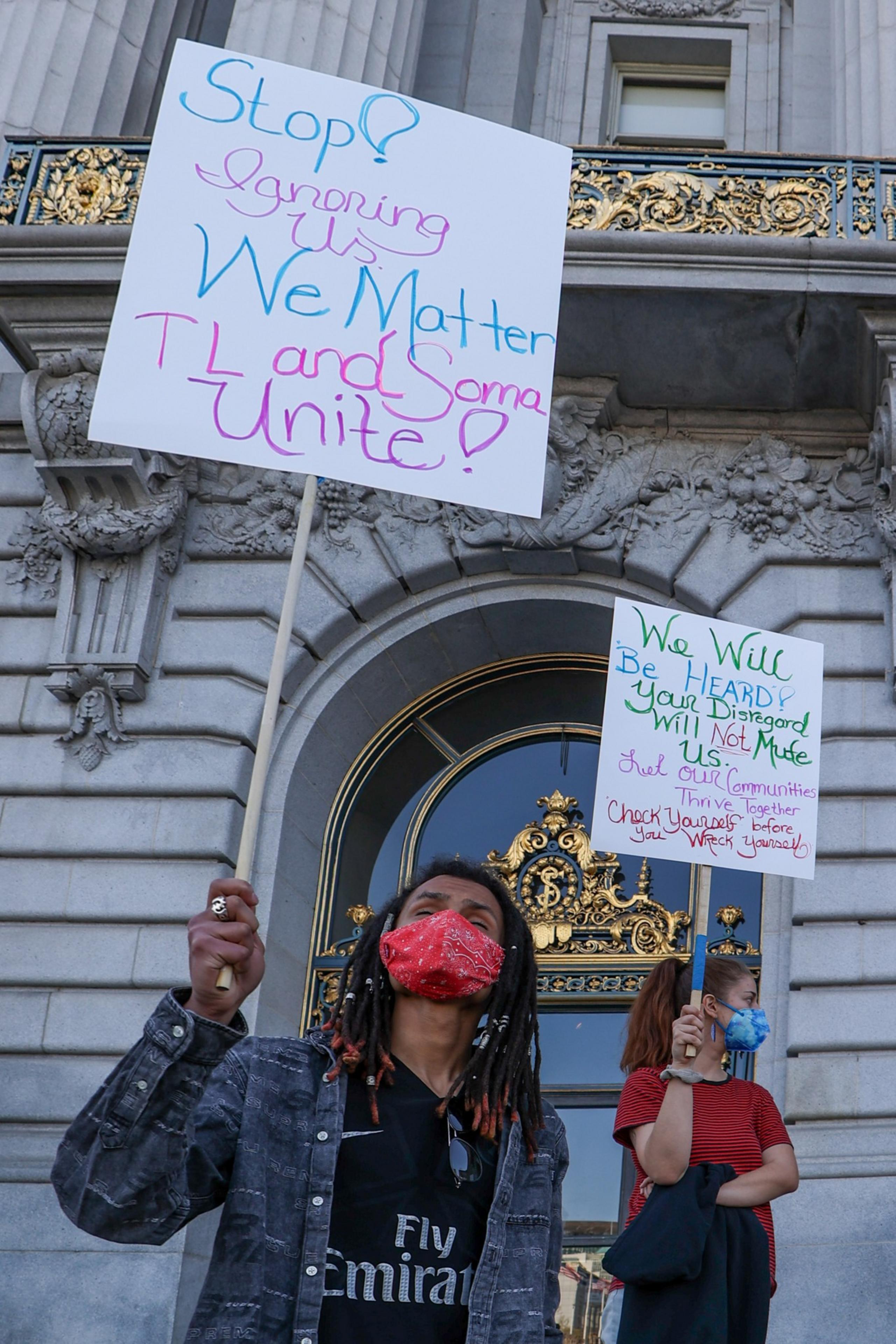 Two masked protesters stand outside a grand building, holding handwritten signs advocating for unity, being heard, and ending disregard.