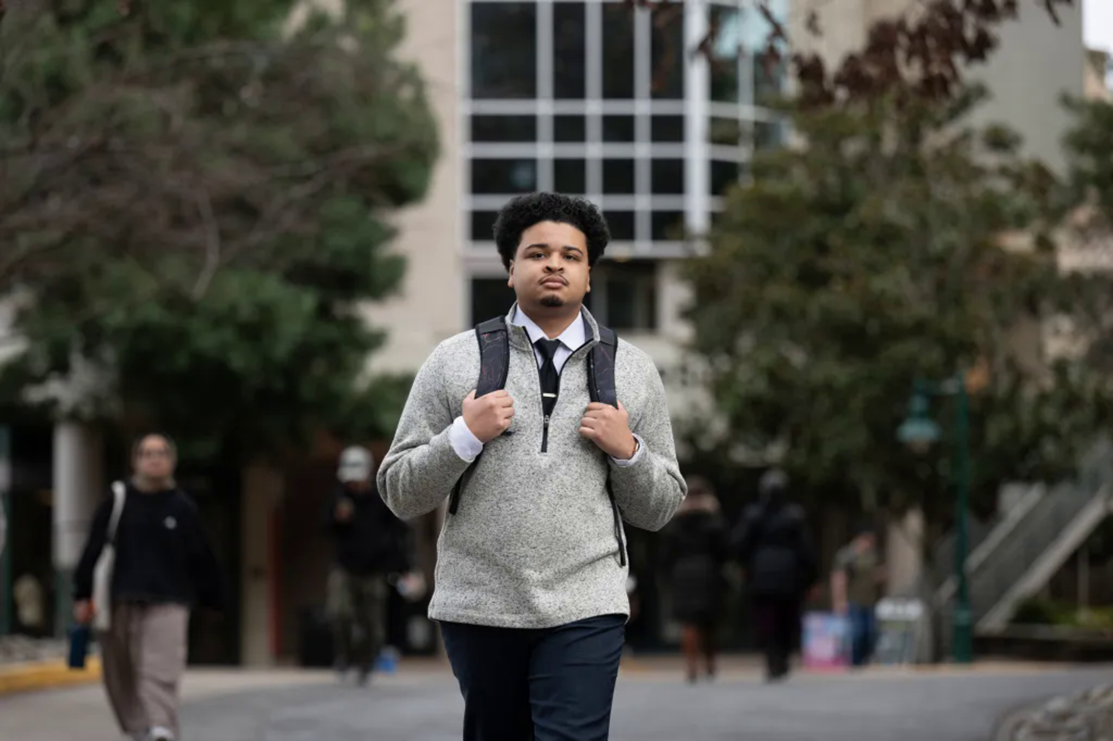A young man walking with a backpack, trees and a building in the background.