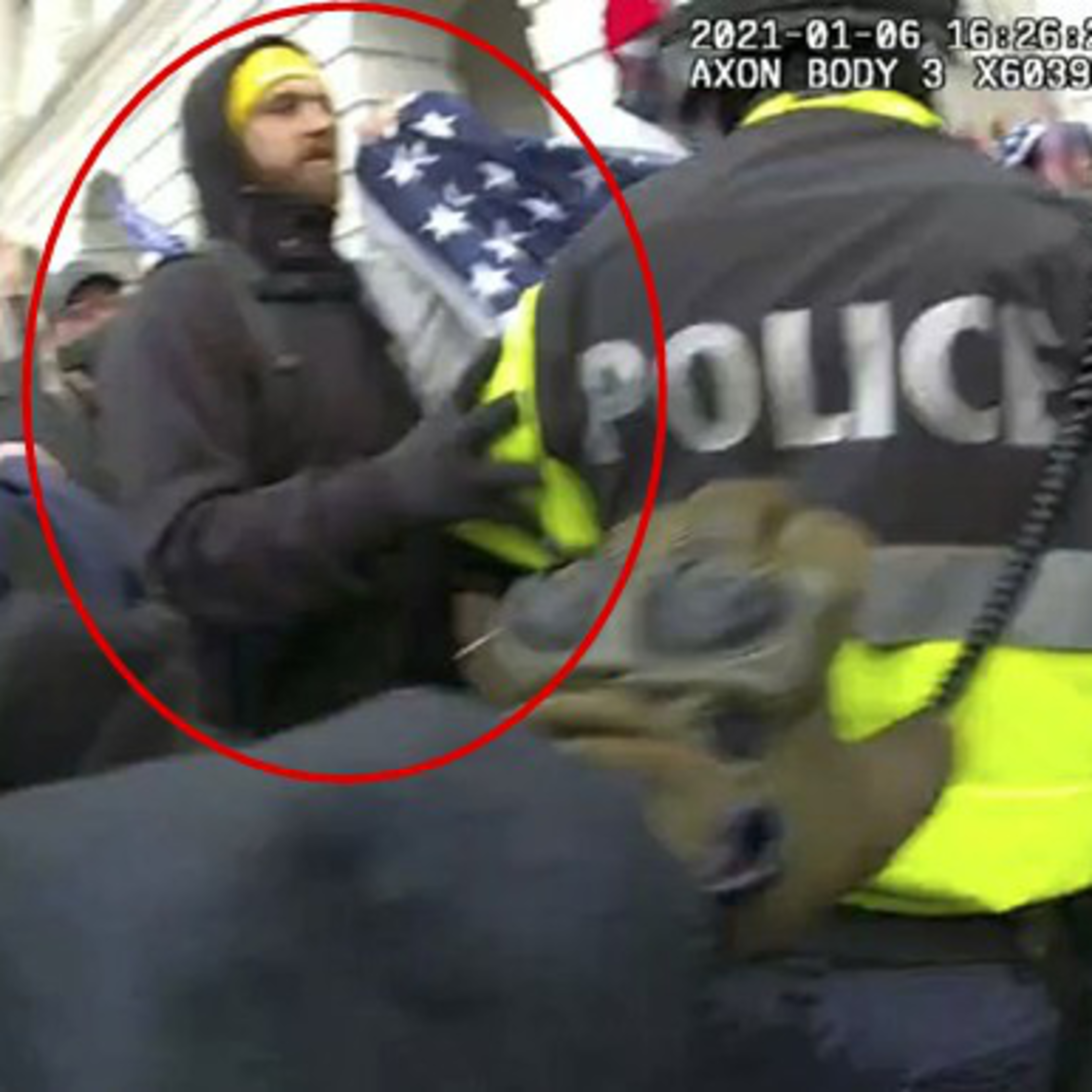 A person in a yellow hat holds a flag beside a police officer in a reflective jacket amidst a crowded scene. The timestamp and "AXON BODY" are visible.