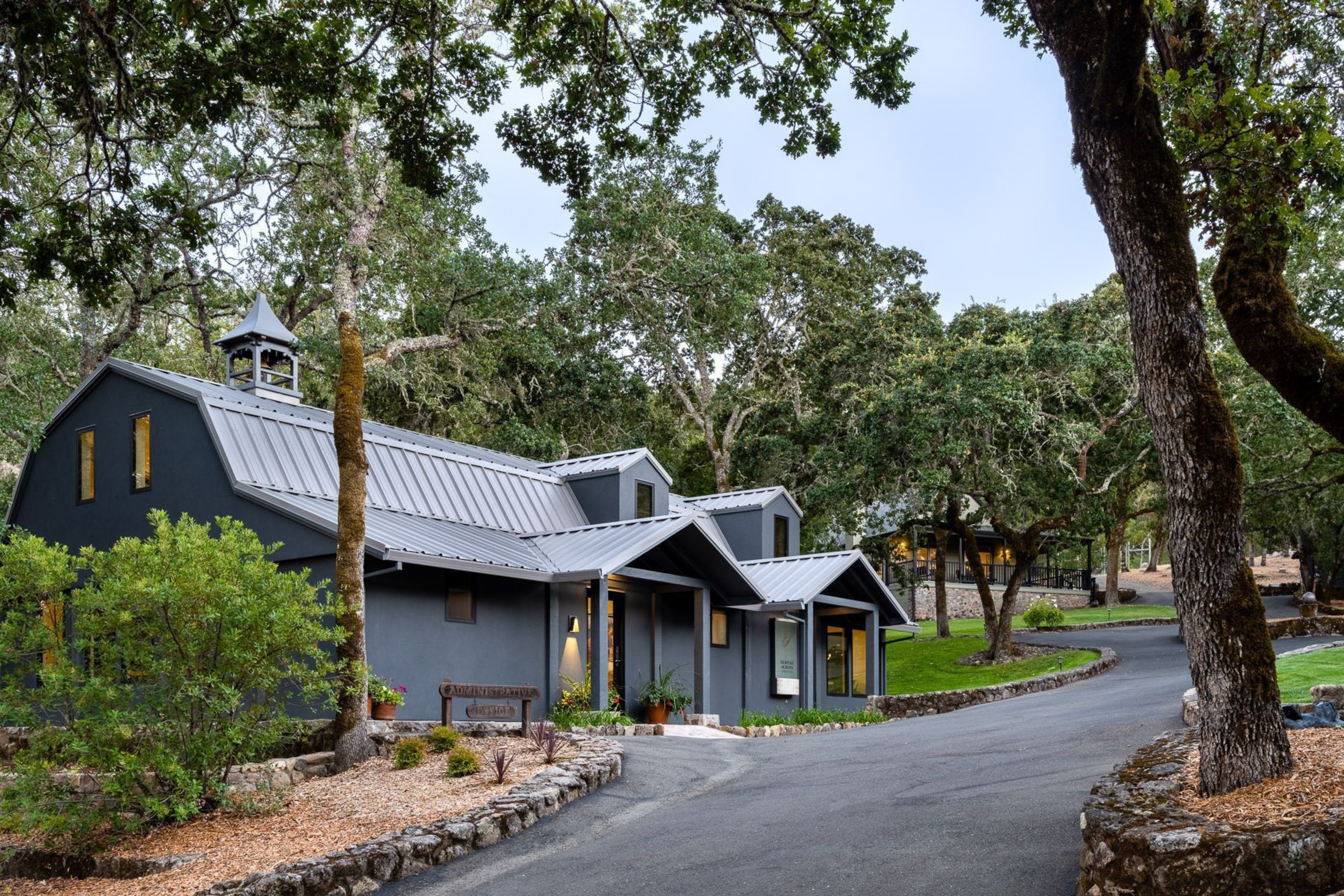 A dark gray barn-style building with a metal roof sits beside a curved paved road, surrounded by trees and greenery in a peaceful natural setting.