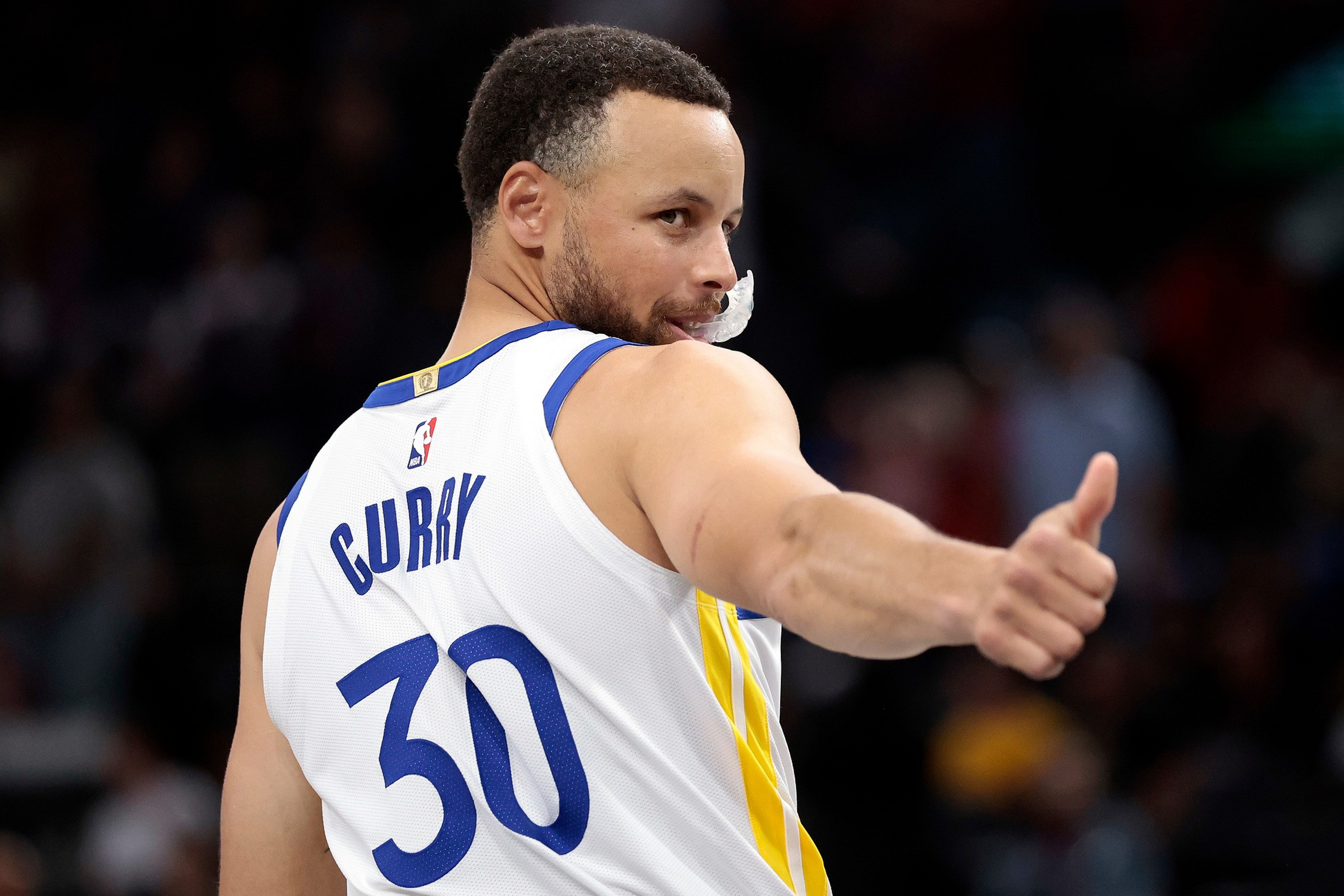 A basketball player wearing a white number 30 jersey with "Curry" on the back looks over his shoulder and gives a thumbs-up.