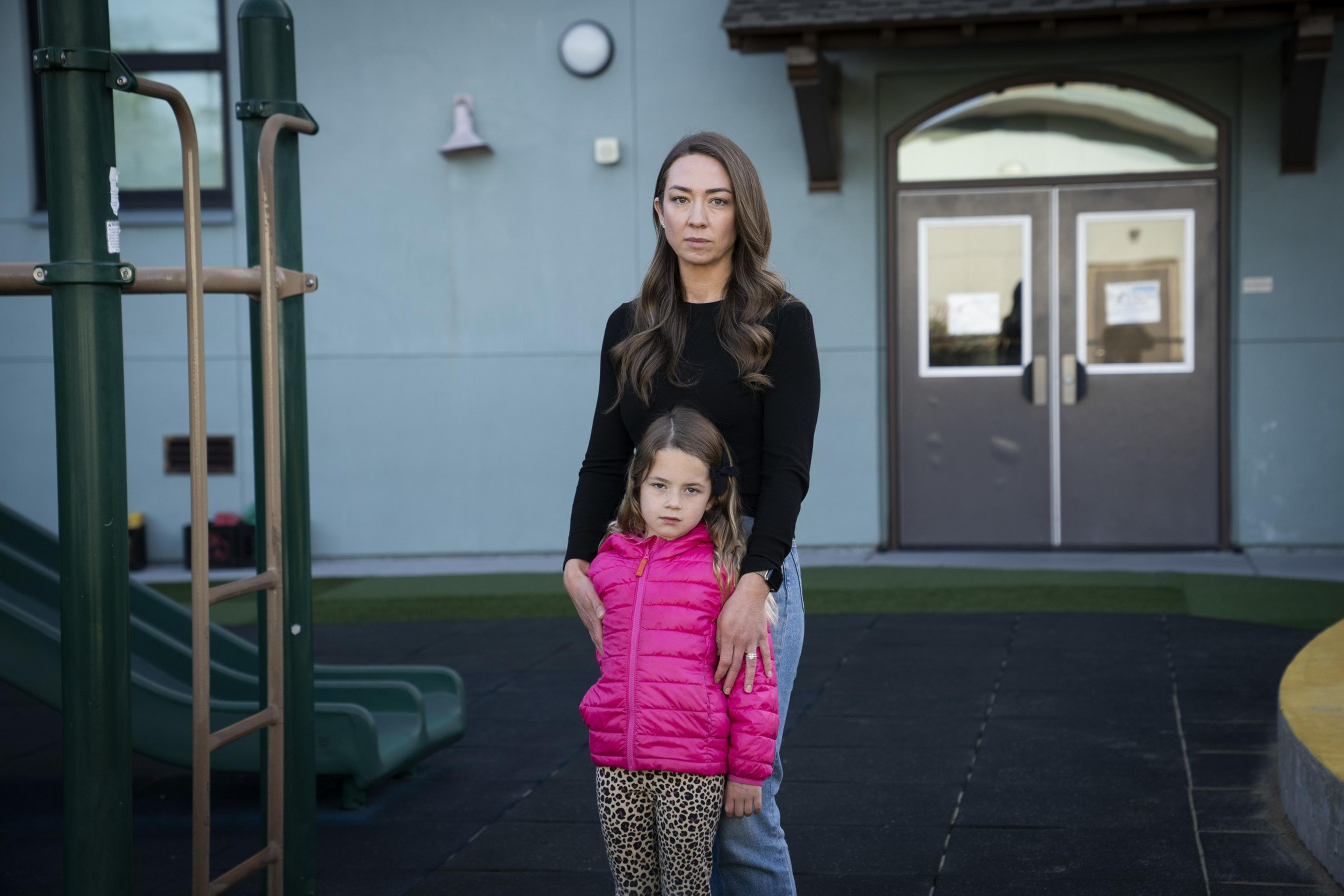 A woman in a black top stands next to a young girl in a bright pink jacket and leopard print leggings. They're in front of a playground and a building entrance.