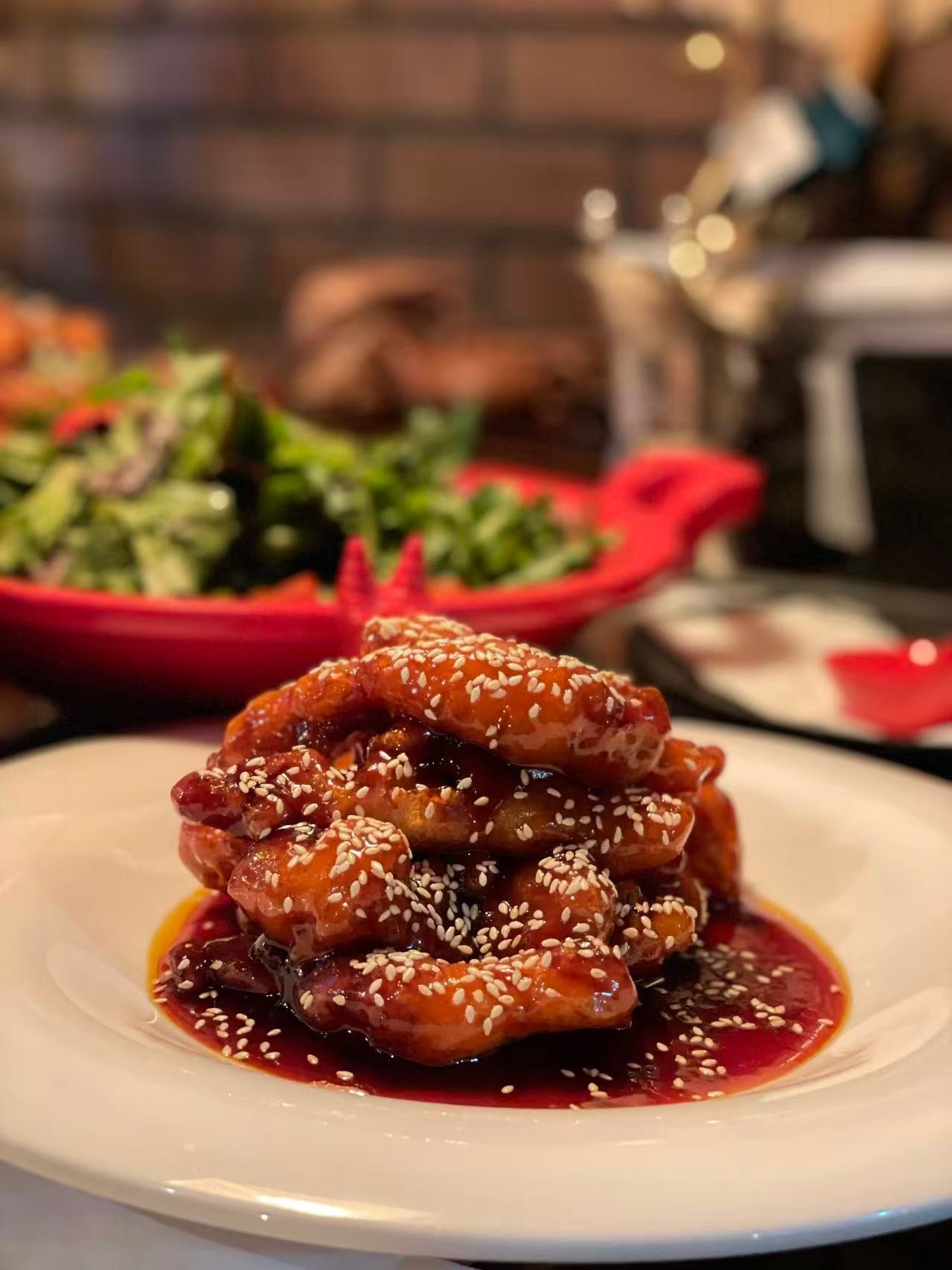 A stack of sesame-coated, glazed chicken wings sits on a white plate with a dark, thick sauce beneath, and a salad on a red dish in the background.