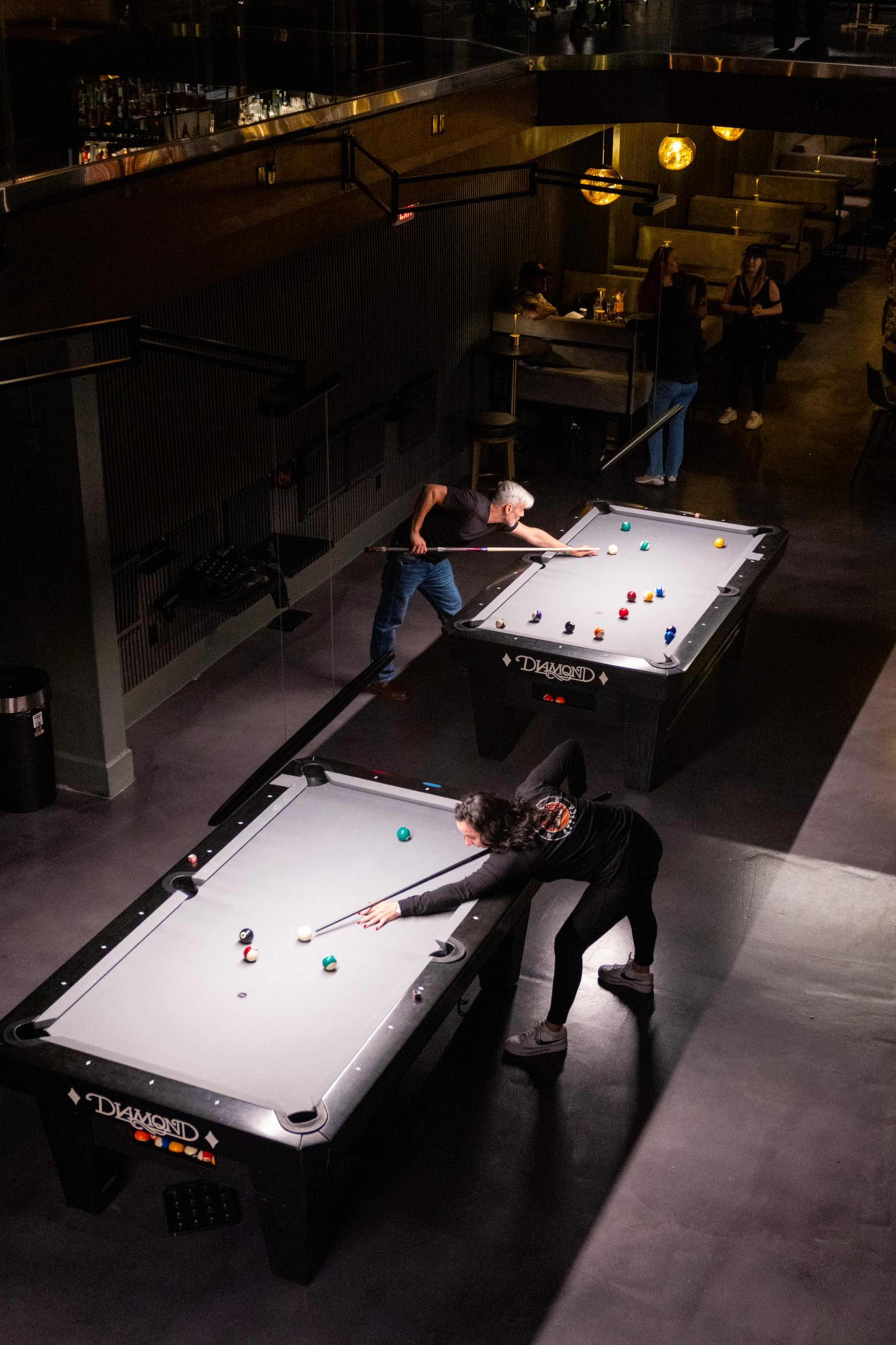 Two people are playing pool on separate tables in a dimly lit room with booths and hanging round lights in the background.