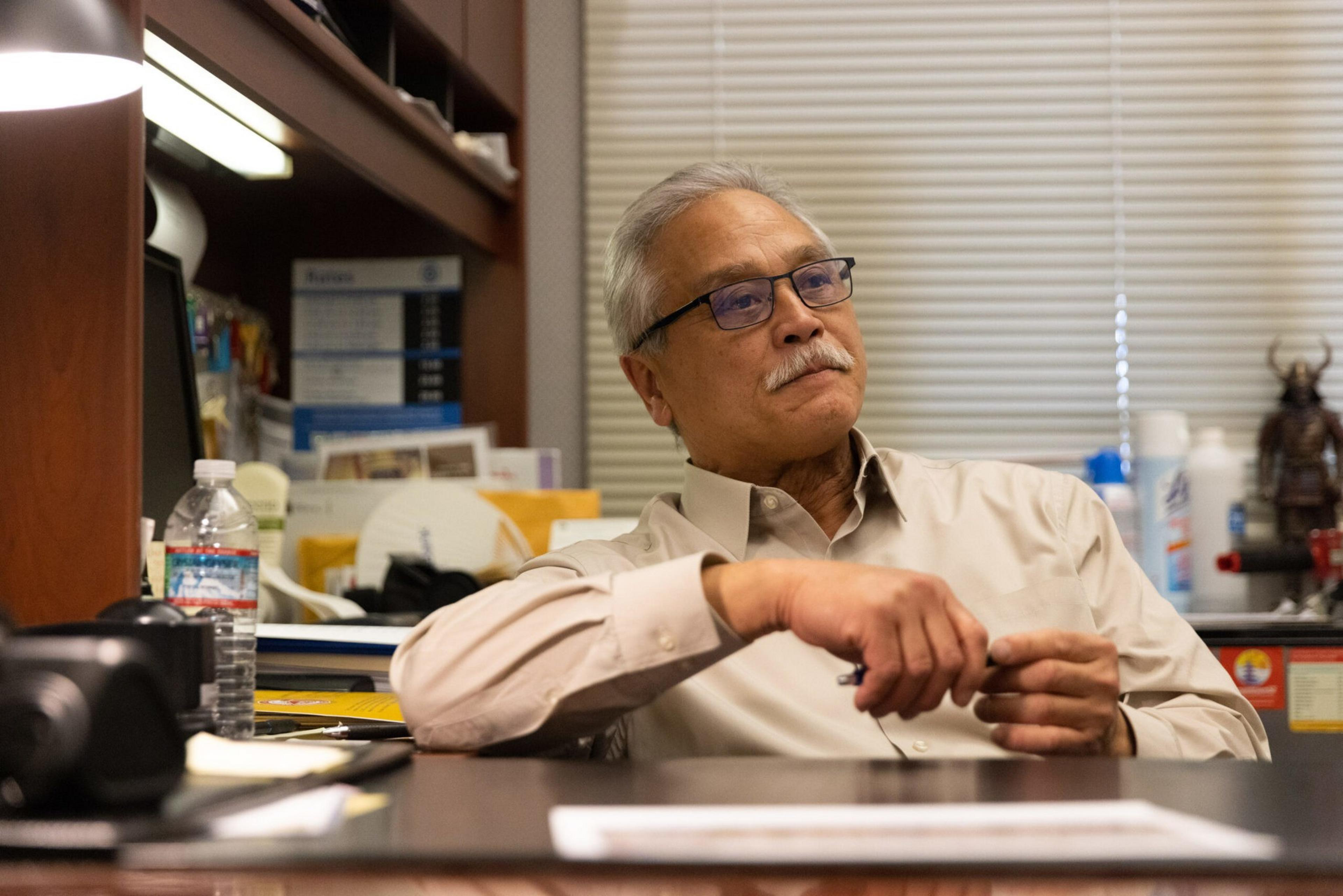 An older man with gray hair and glasses sits at a cluttered desk in an office, holding a pen and looking thoughtfully to the side.