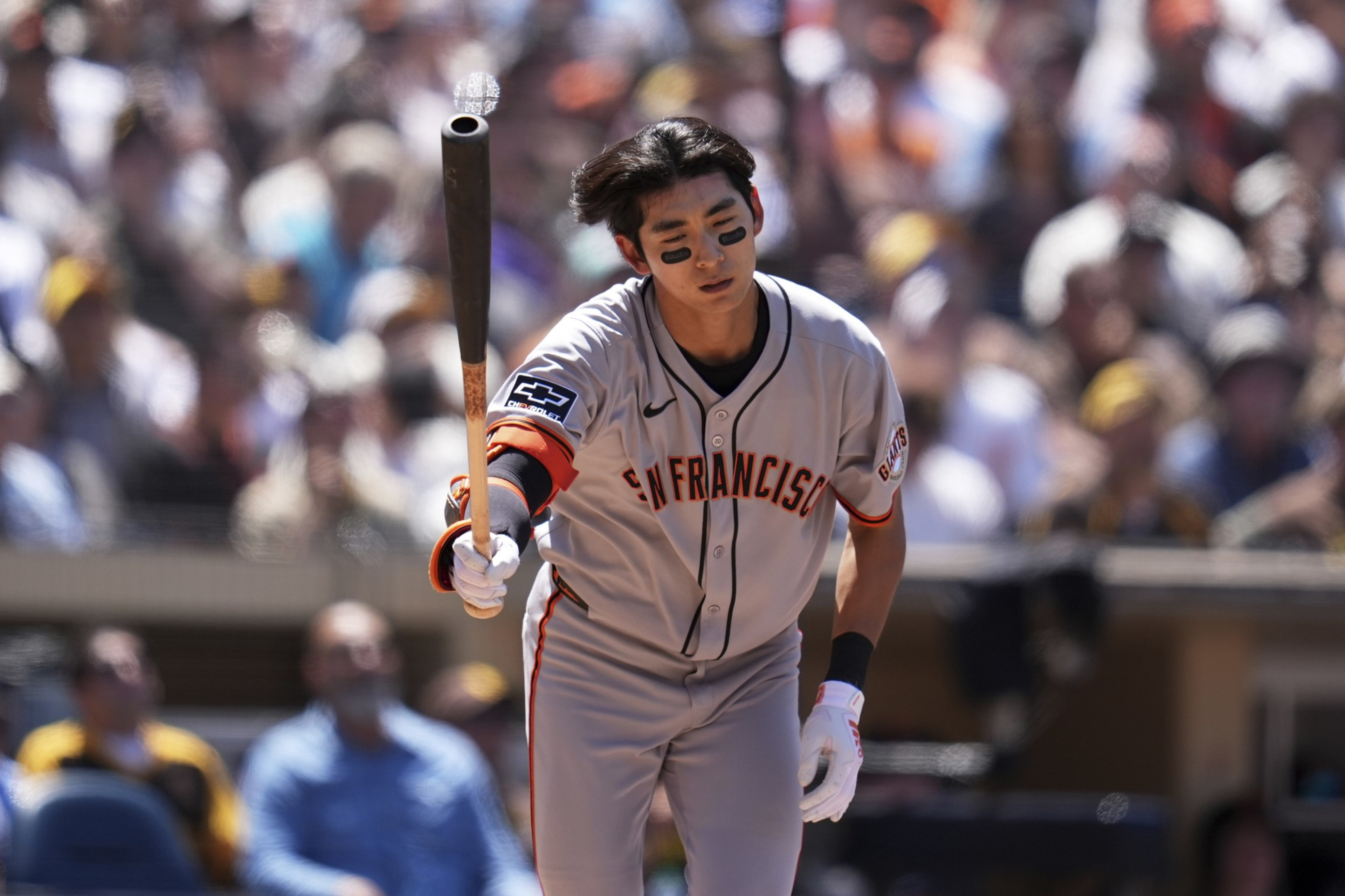 A baseball player in a gray uniform with "San Francisco" written on it holds a bat, with a focused expression. A blurred crowd is in the background.